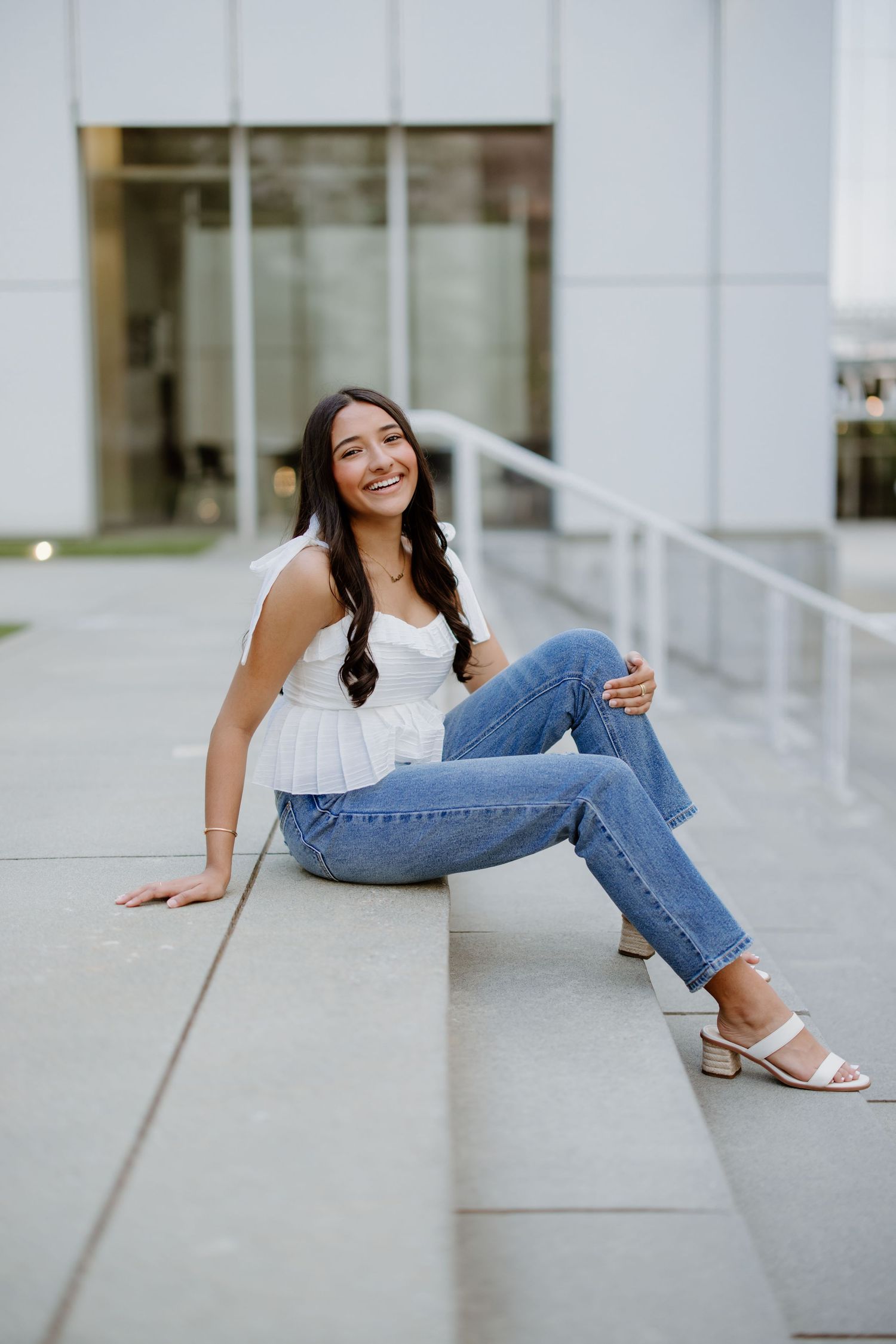 Person in casual summer outfit with white top and ripped jeans sits on concrete steps with a bright smile.