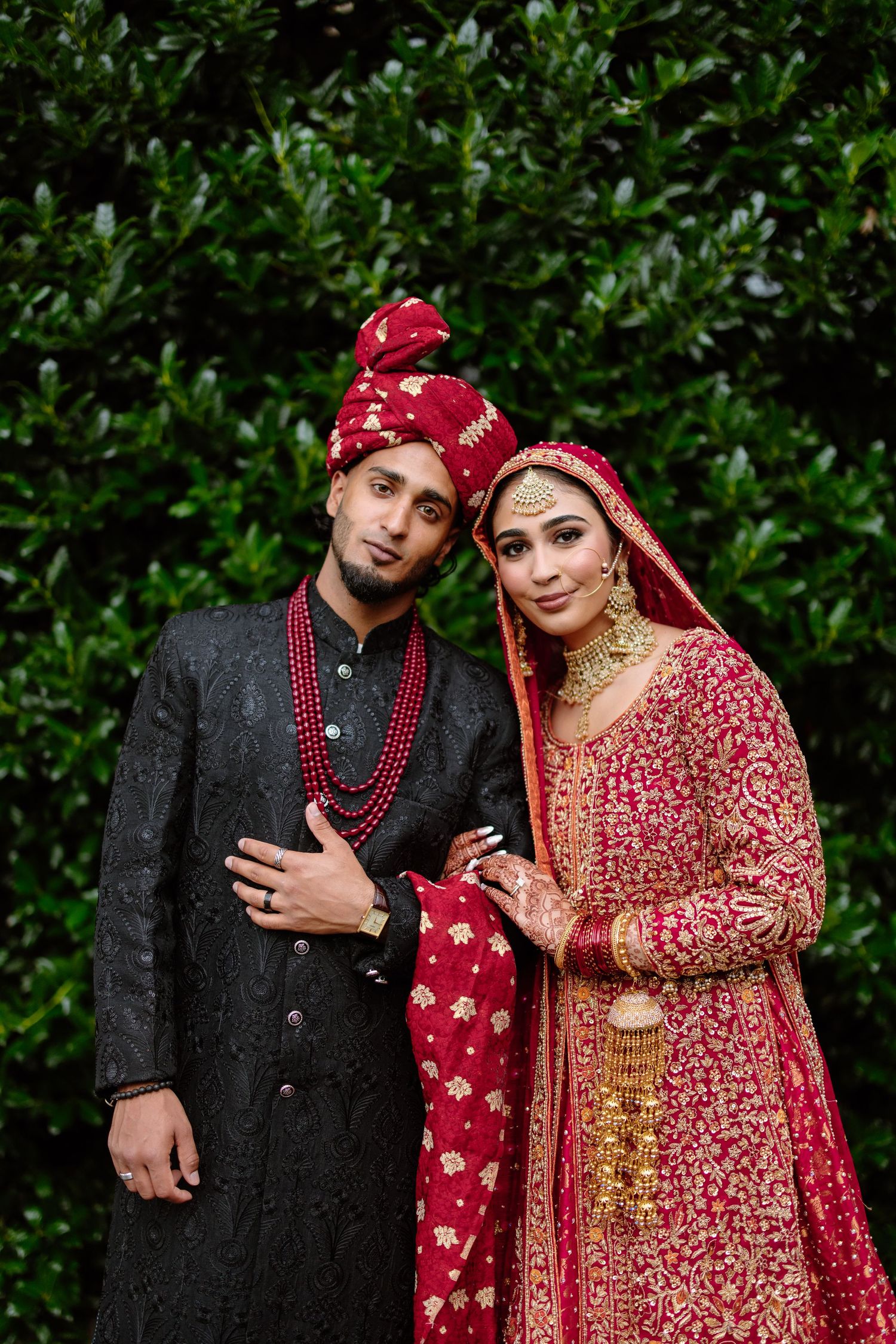 A couple in traditional South Asian wedding attire pose together against a green hedge background.