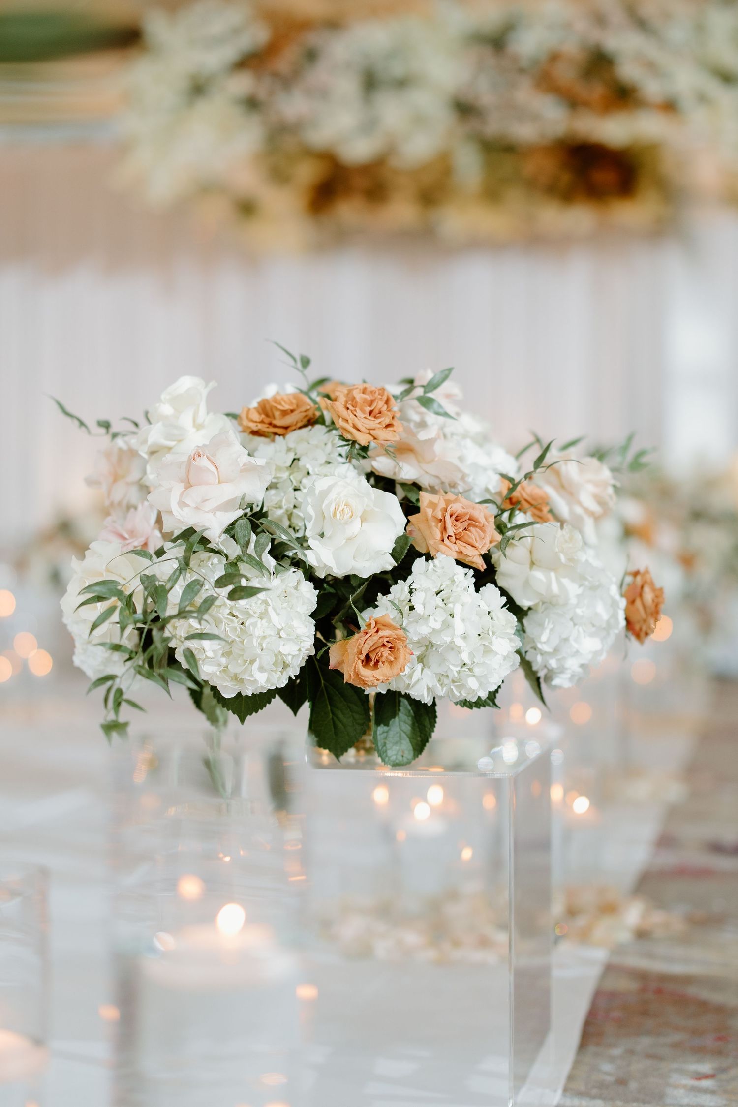 Elegant wedding centerpiece with white hydrangeas and peach roses on clear acrylic stand with glowing candles.
