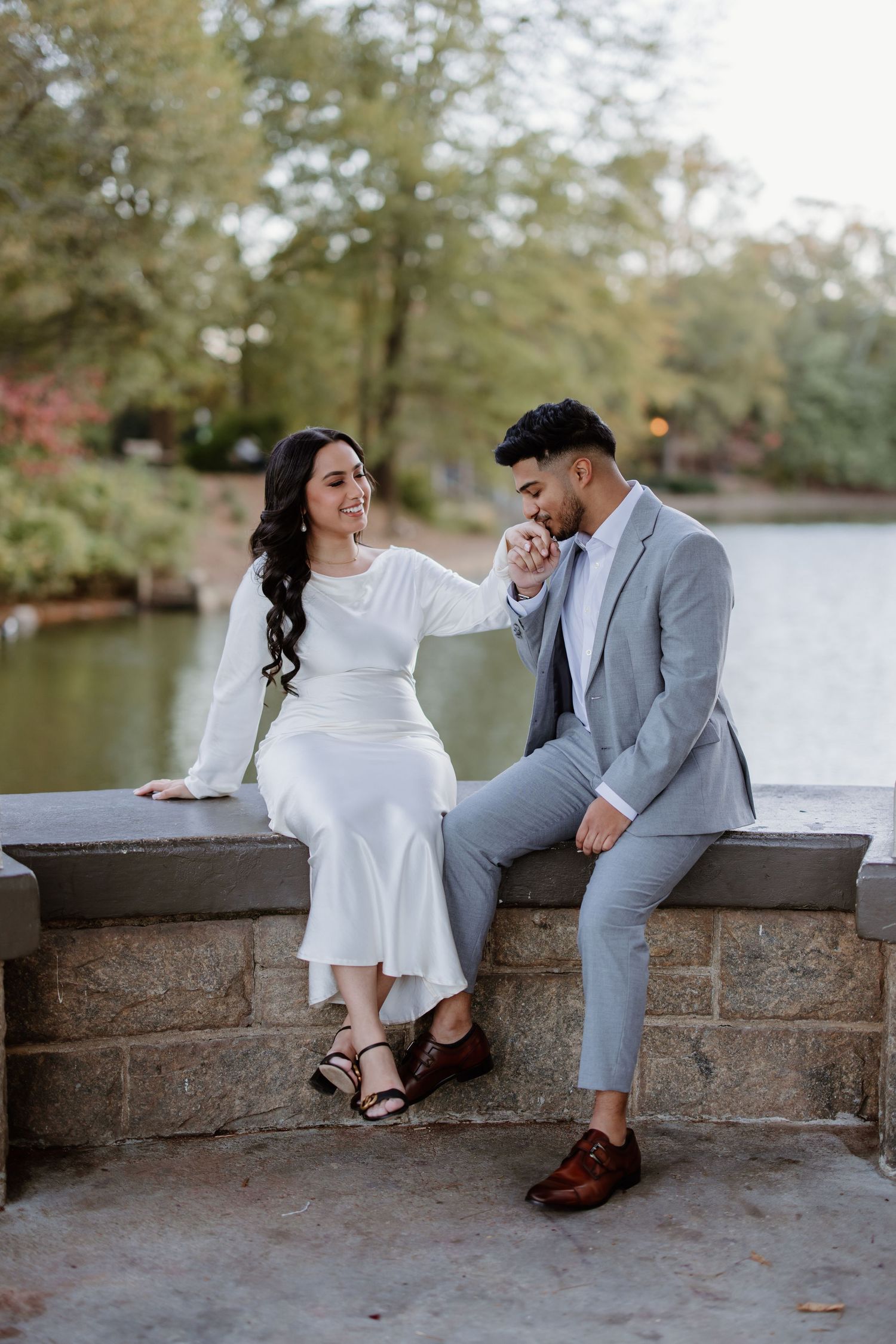 A couple sharing romantic moments while sitting on a stone ledge by a tranquil lake in an autumn setting.