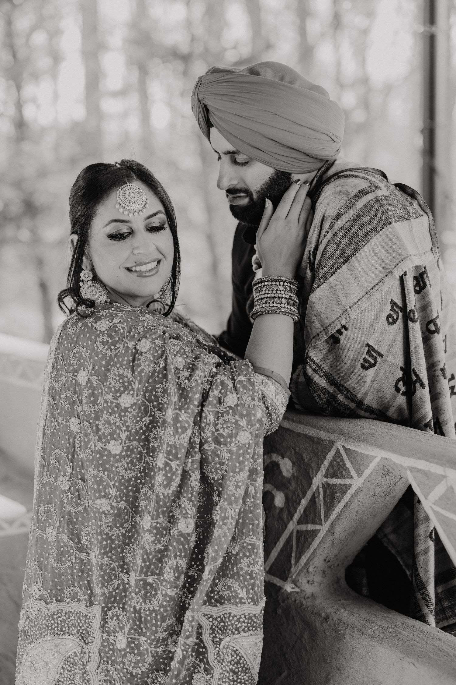 Black and white portrait of a couple in traditional Punjabi wedding attire sharing an intimate moment outdoors.