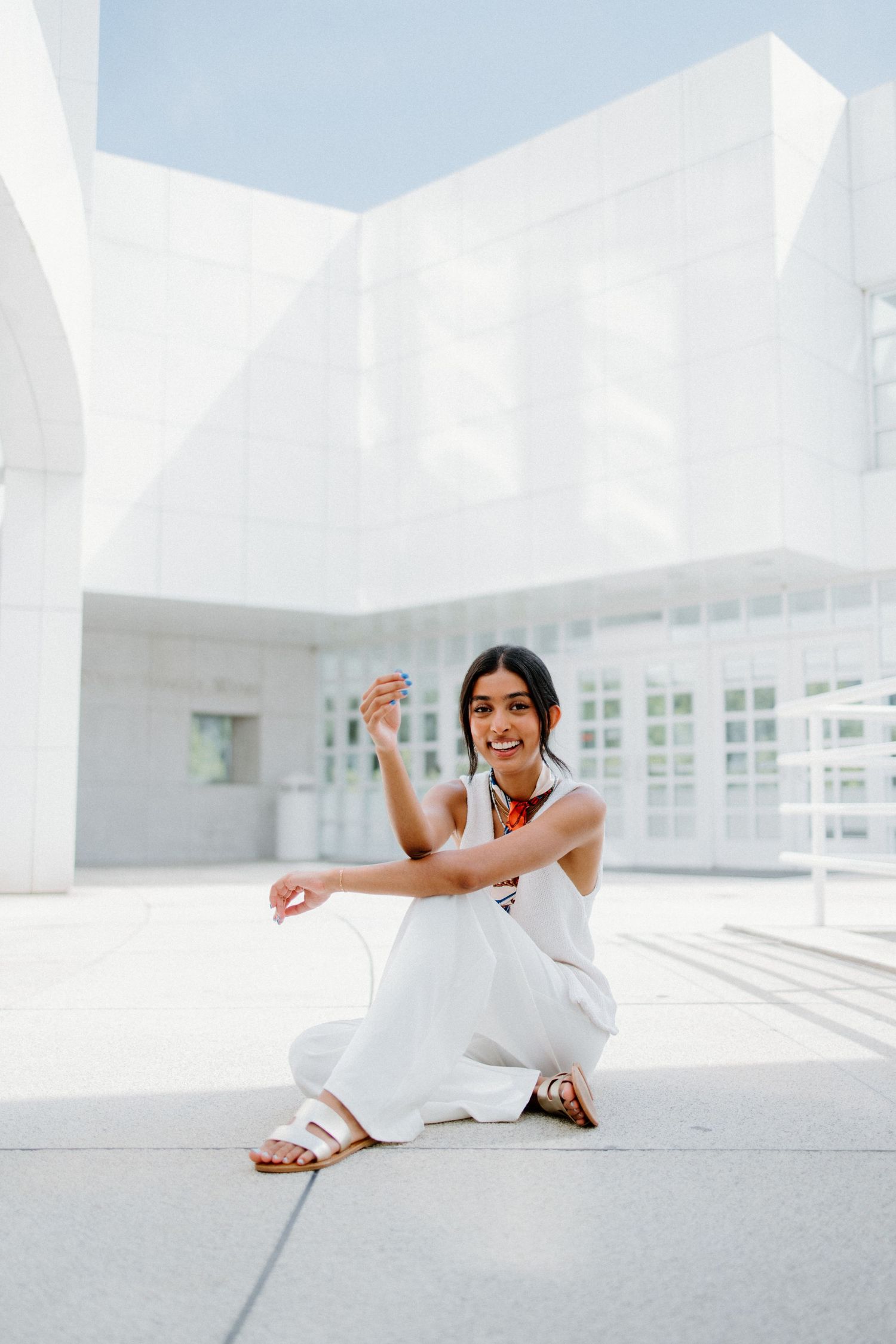 Someone in an all-white outfit sits and smiles in front of a modern white architectural building on a sunny day.