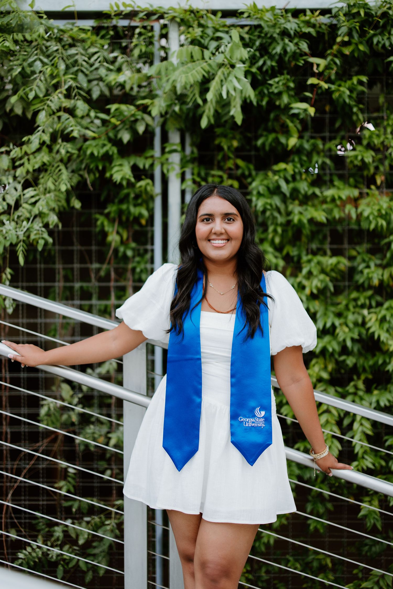 Graduate in white dress and blue graduation stole poses outdoors against a green leafy backdrop.