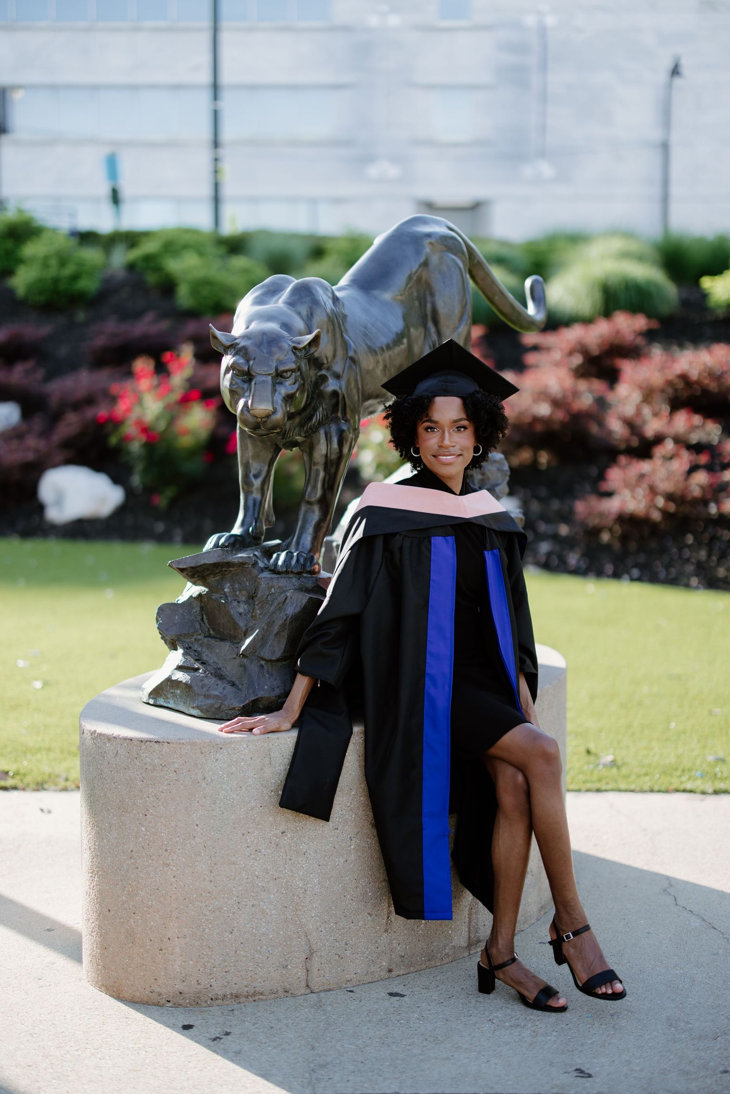 Graduate poses in academic regalia next to a metallic panther statue on a stone pedestal on a sunny day.