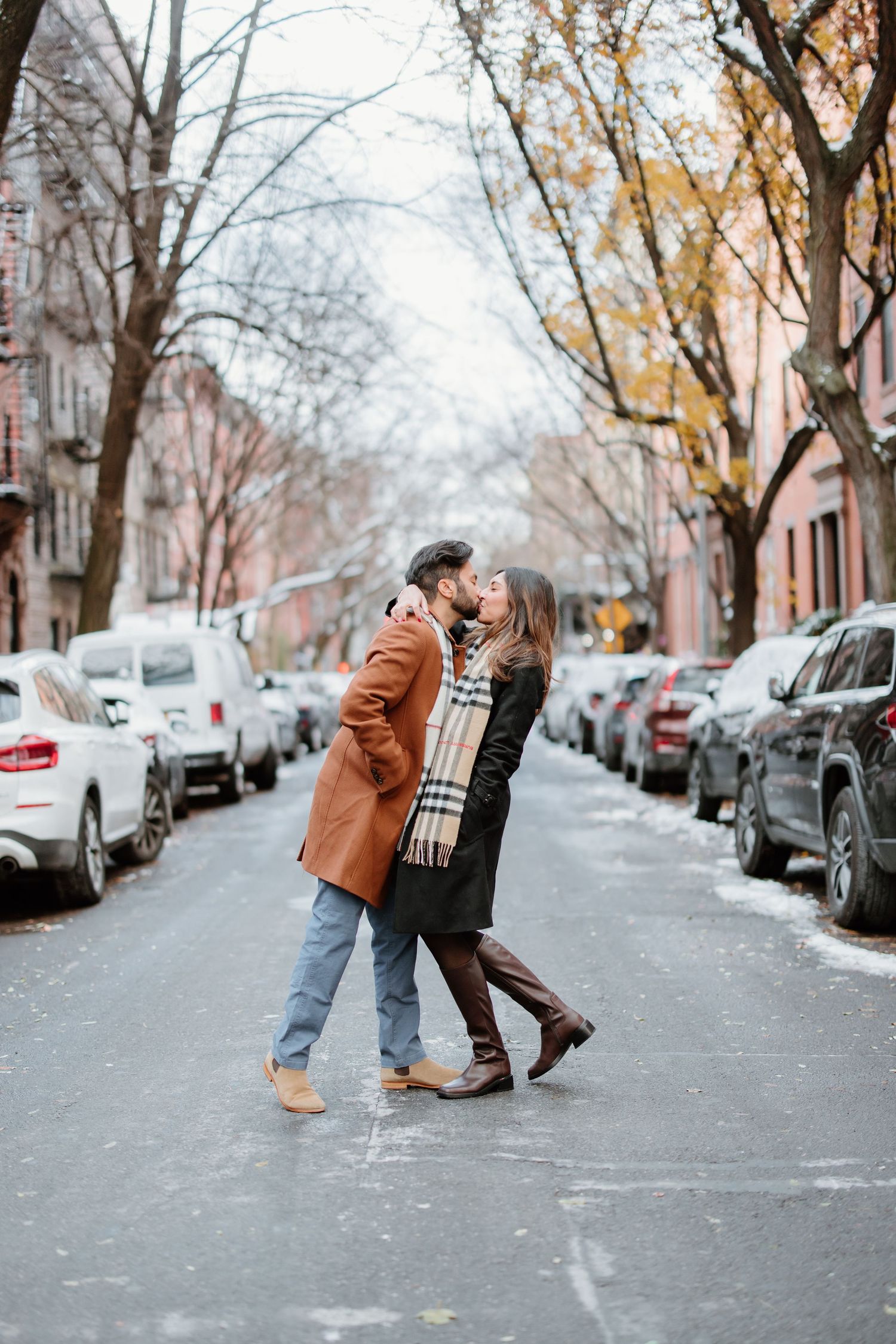 A couple plays and dances together on a snow-lined city street with parked cars and bare winter trees.
