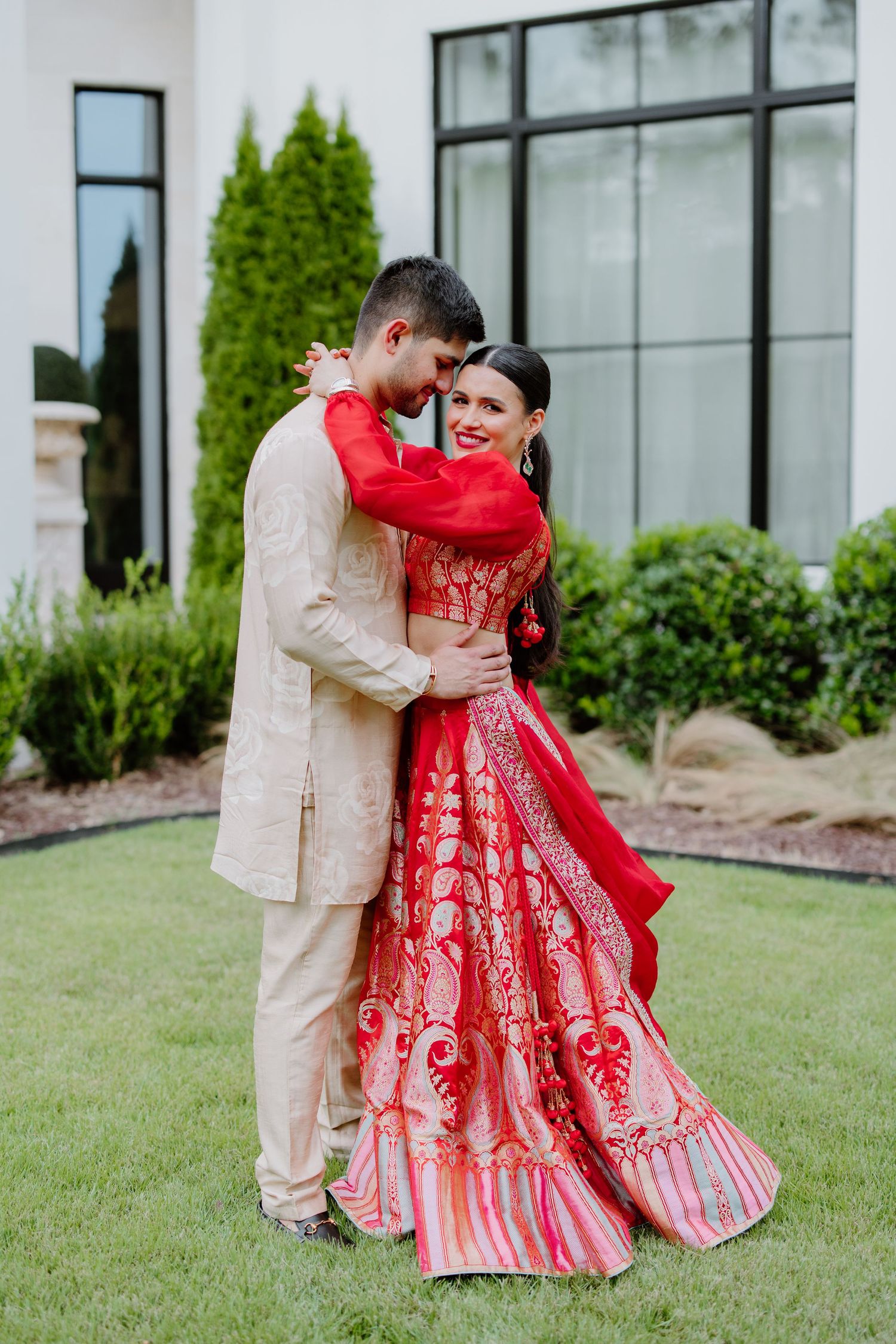 A couple in traditional South Asian wedding attire embrace on a lawn, with the bride wearing a red embroidered lehenga.