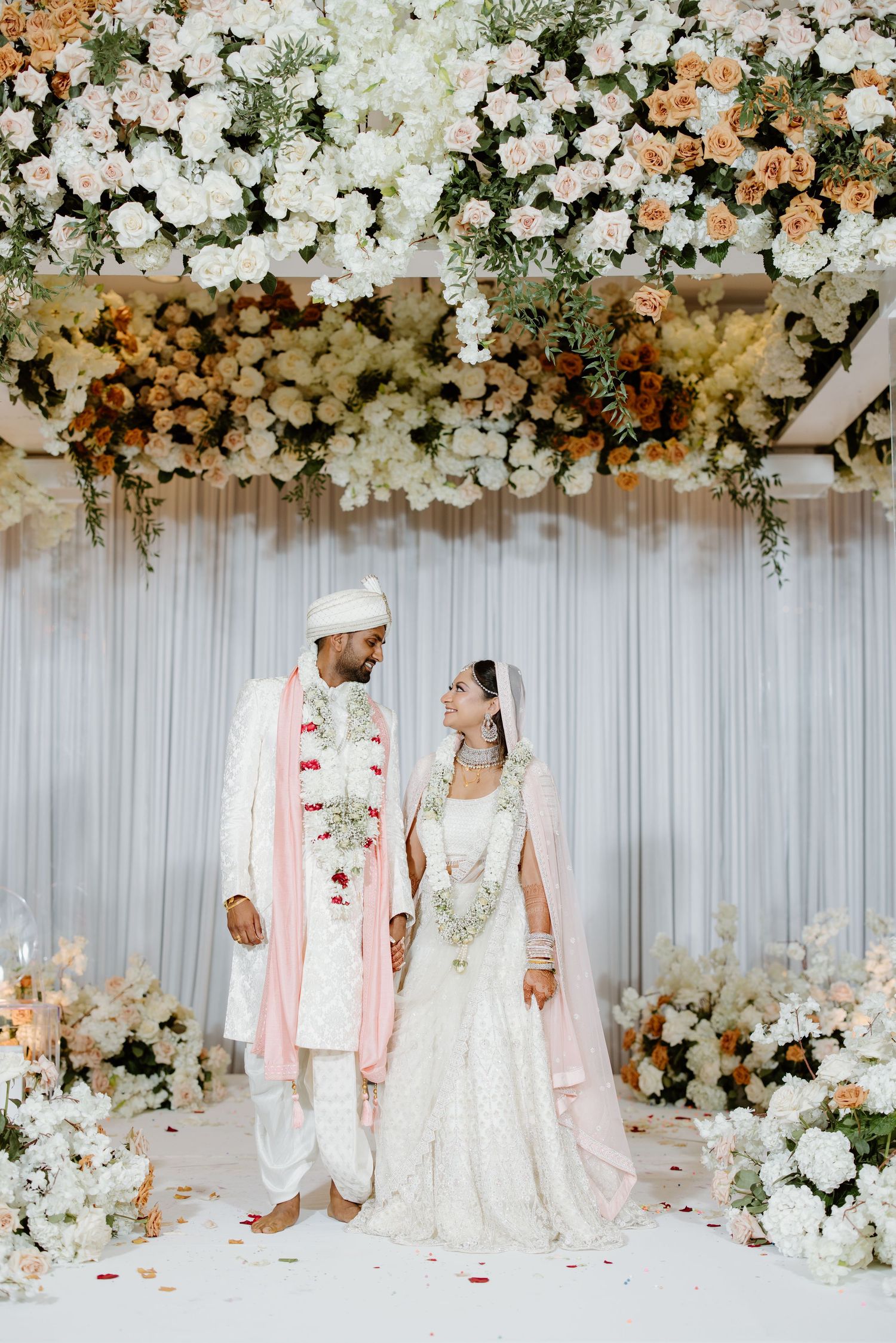 Traditional Indian wedding ceremony setup with elegant white and peach floral mandap decorations against a white backdrop.