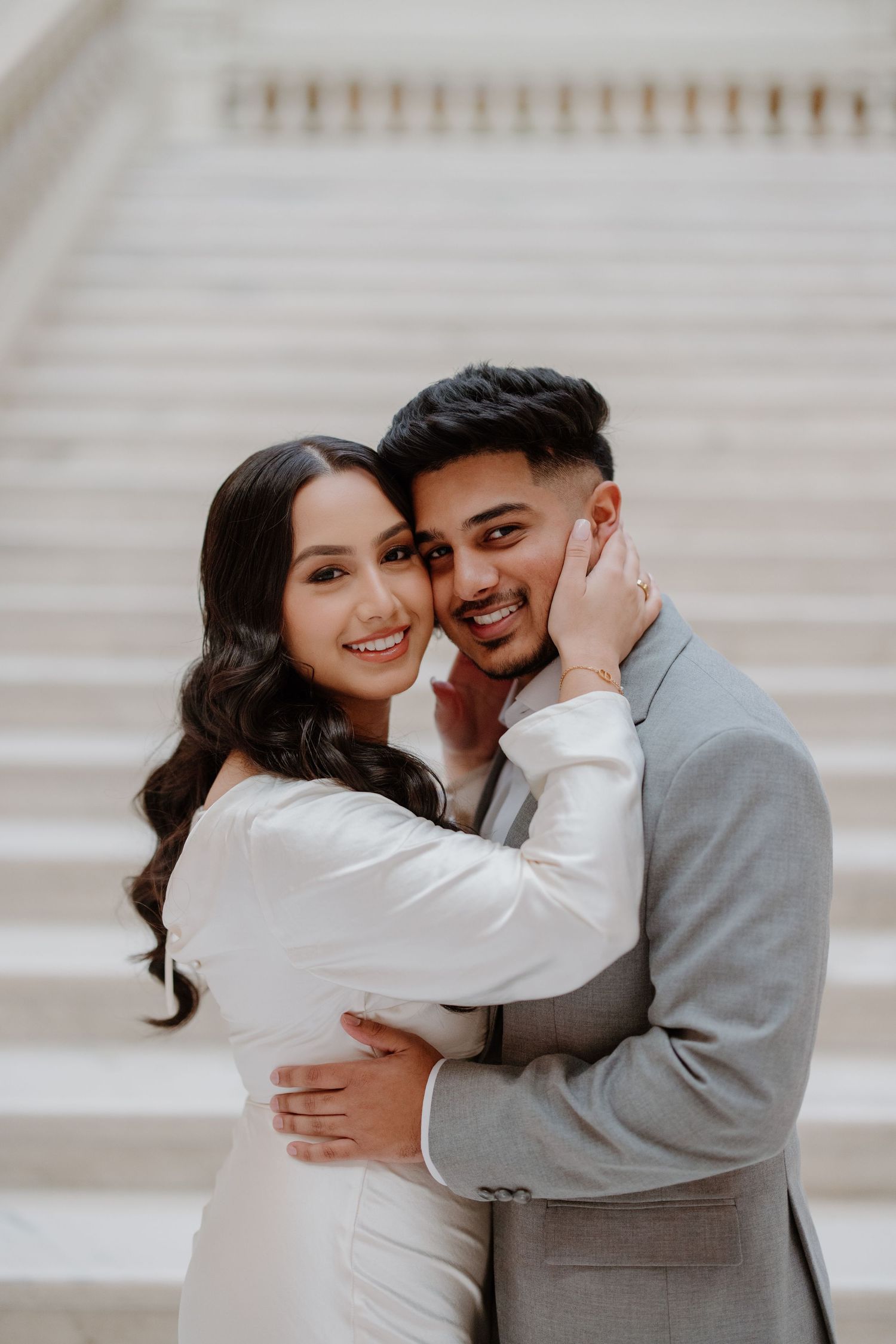 A couple in coordinating white and grey outfits share tender embraces and playful moments on a pale staircase.