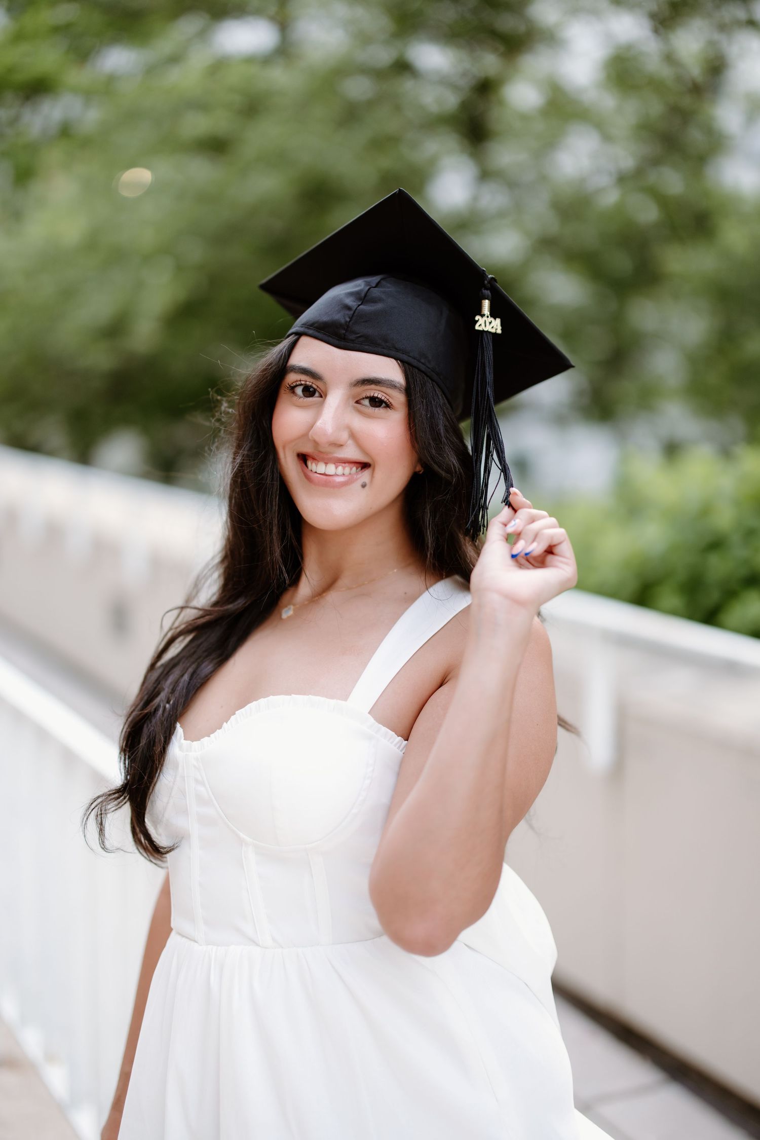 A graduate in white dress and black cap poses with joyful expressions during an outdoor graduation photoshoot.