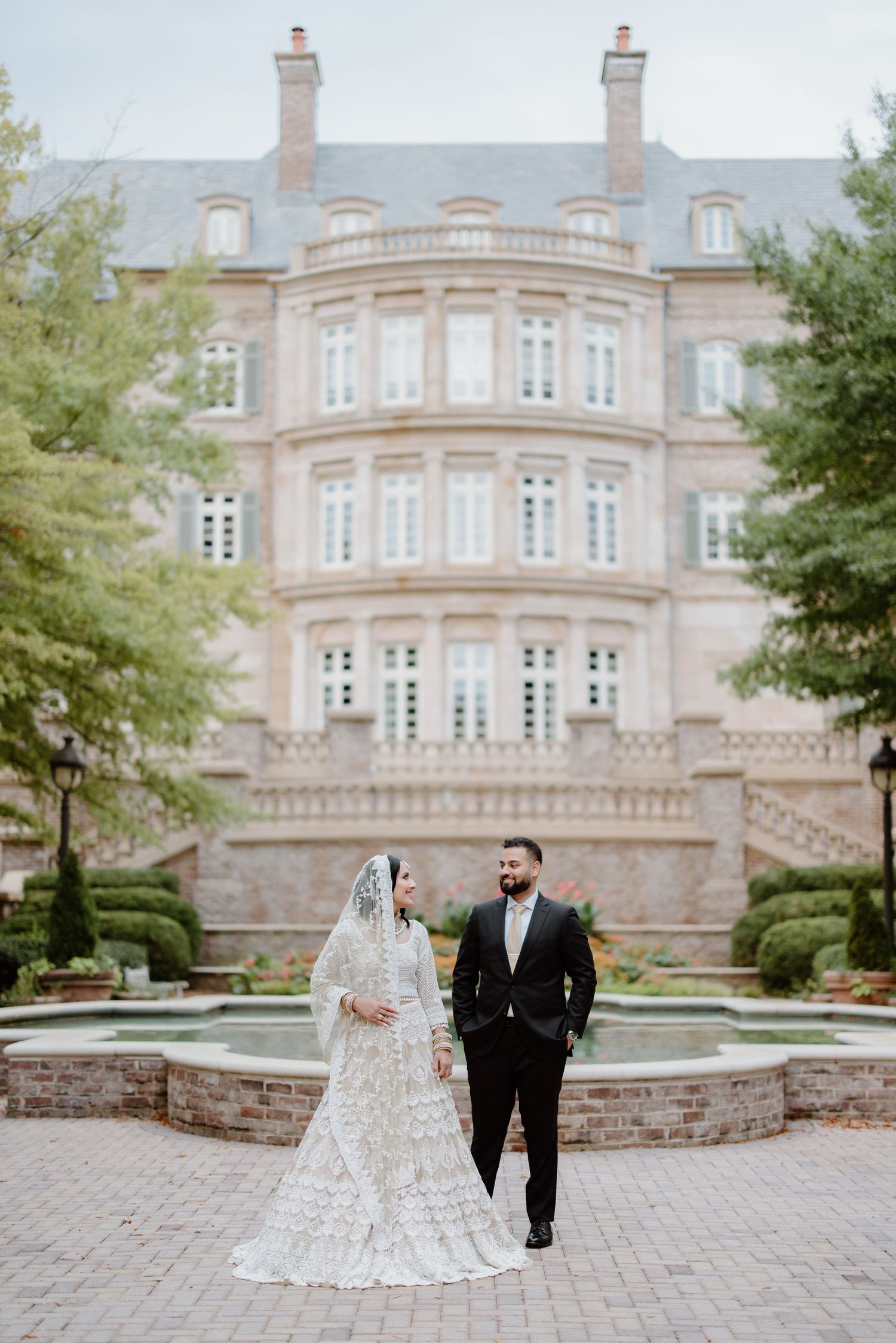 A bridal couple walks and poses along a grand estate entrance with stately architecture and landscaping in the background.