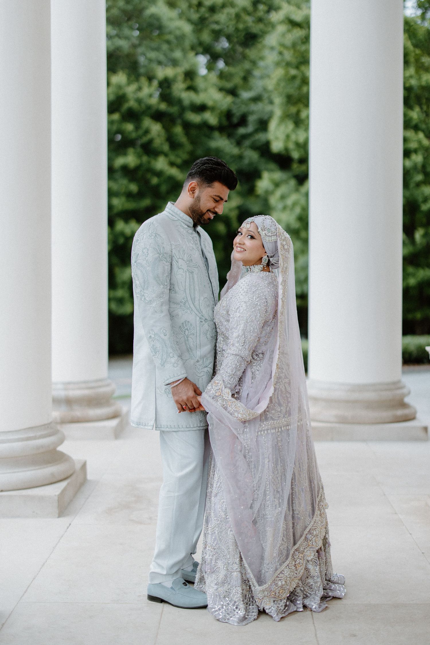 A wedding couple in matching white outfits stands lovingly between classical columns in an outdoor setting.