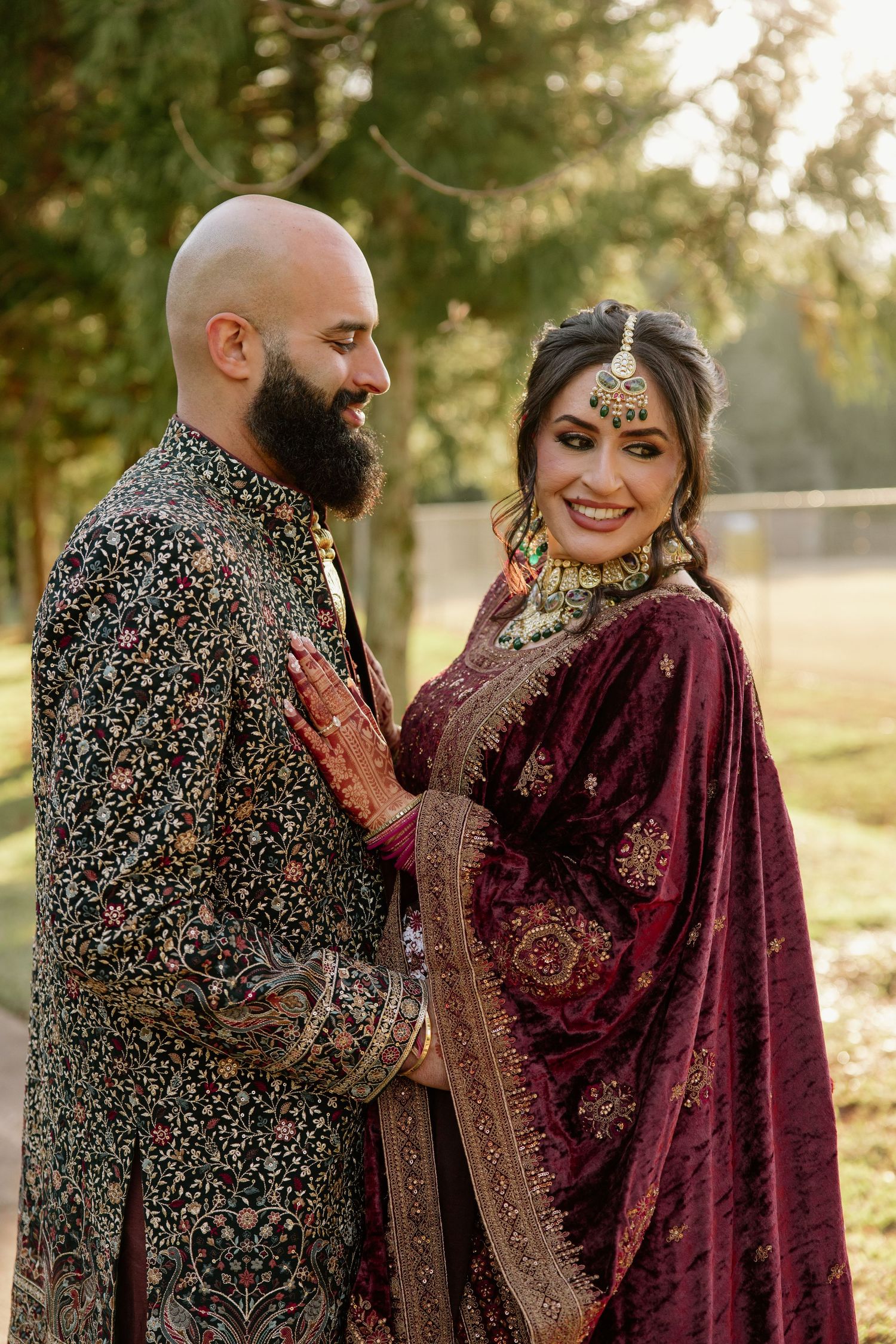 South Asian couple in traditional wedding attire, bride in burgundy lehenga with gold jewelry, groom in floral sherwani.