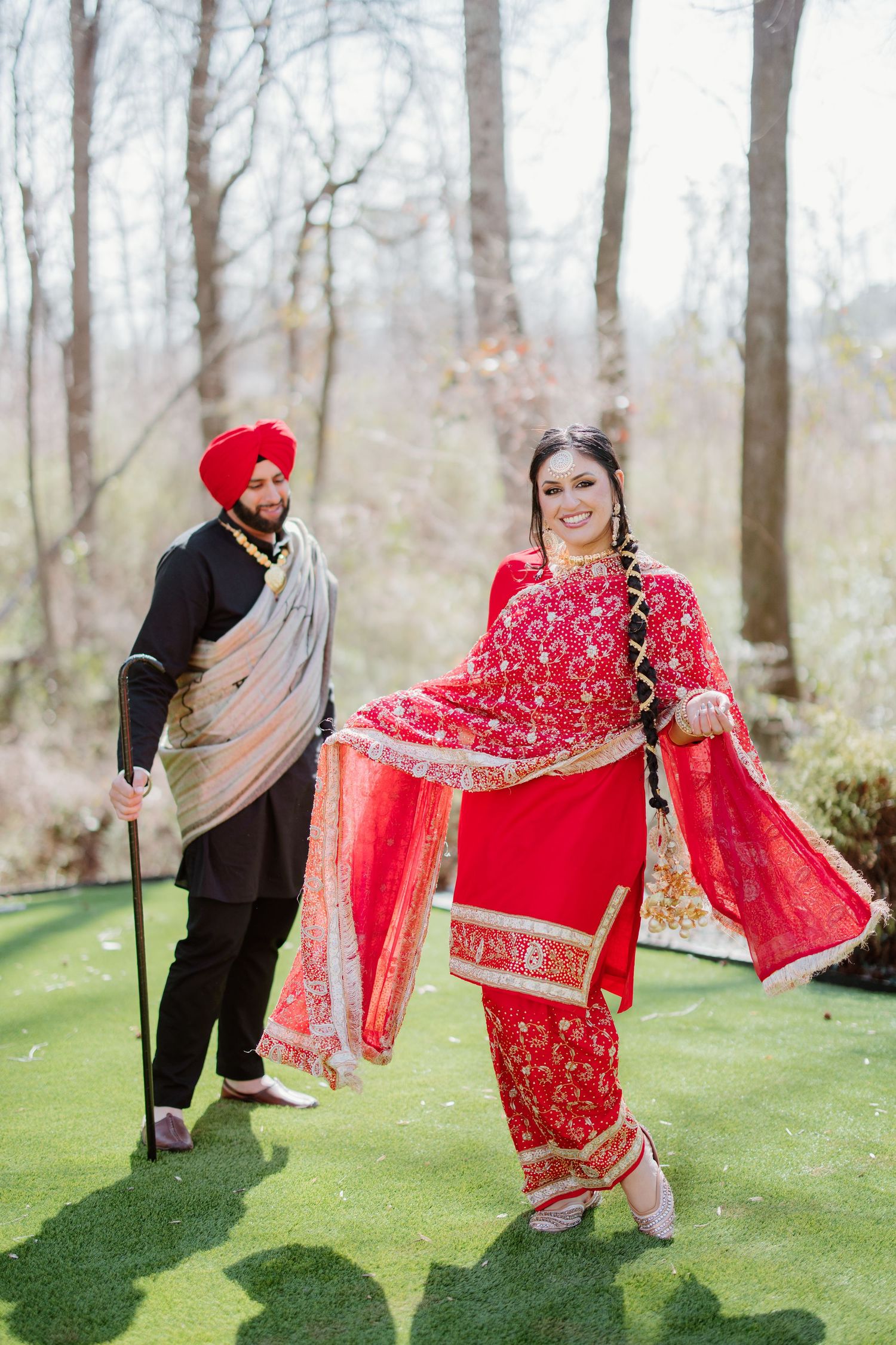A couple in traditional Punjabi wedding attire poses in a garden setting with flowing red and gold garments.