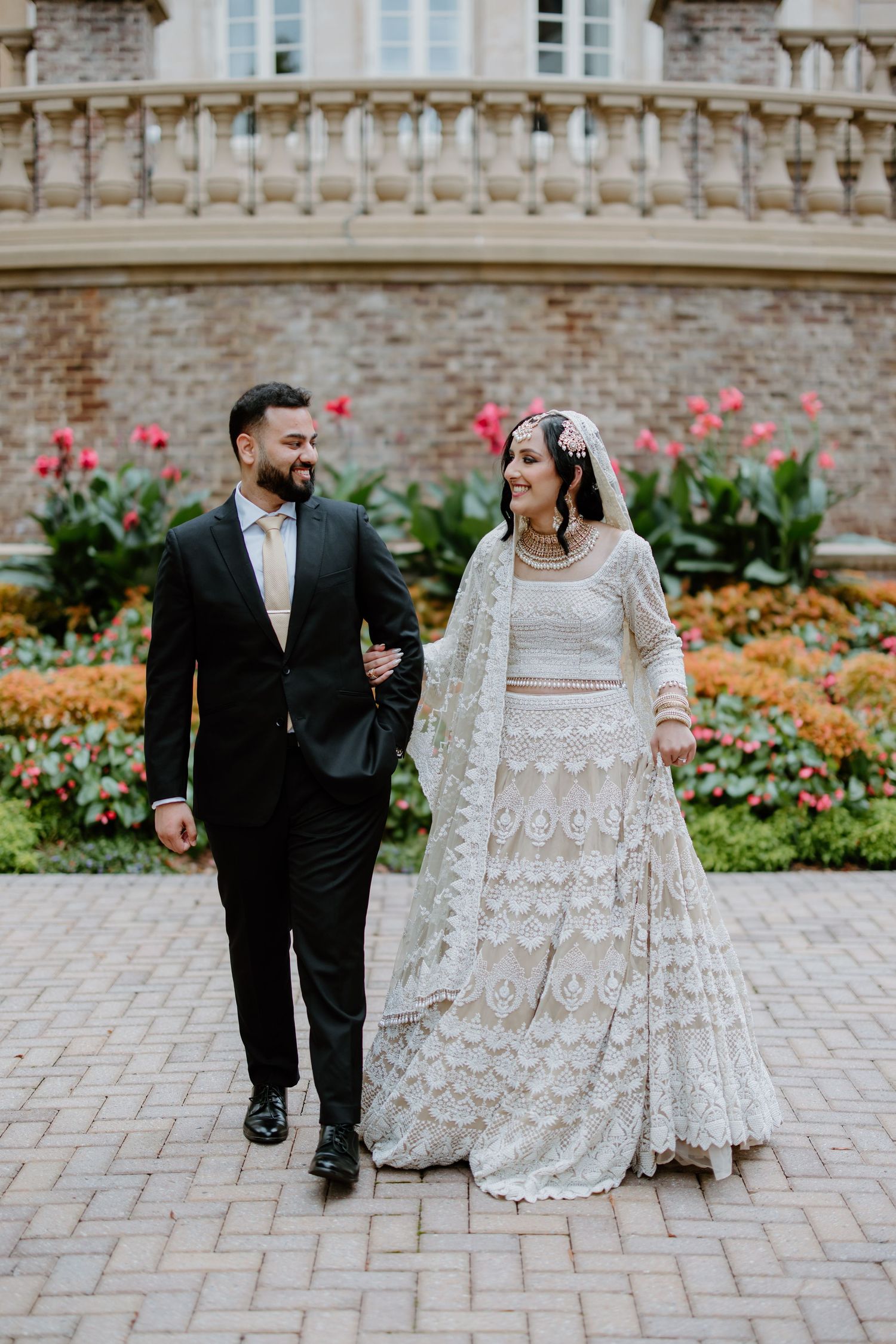 A couple strolls along a paved garden path beside a historic stone building with blooming flowers.