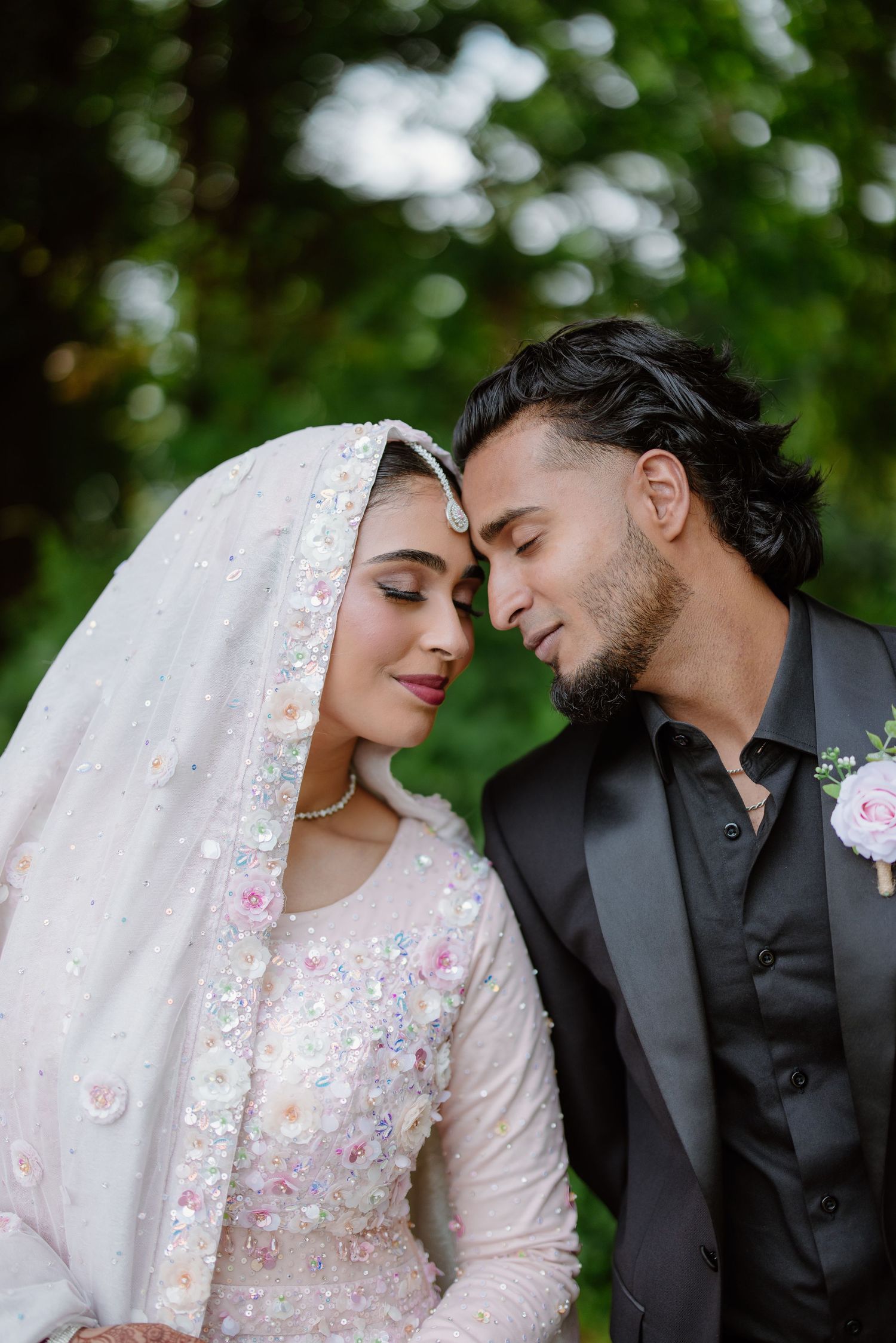 A romantic wedding portrait features a couple in traditional South Asian bridal attire against a green outdoor background.