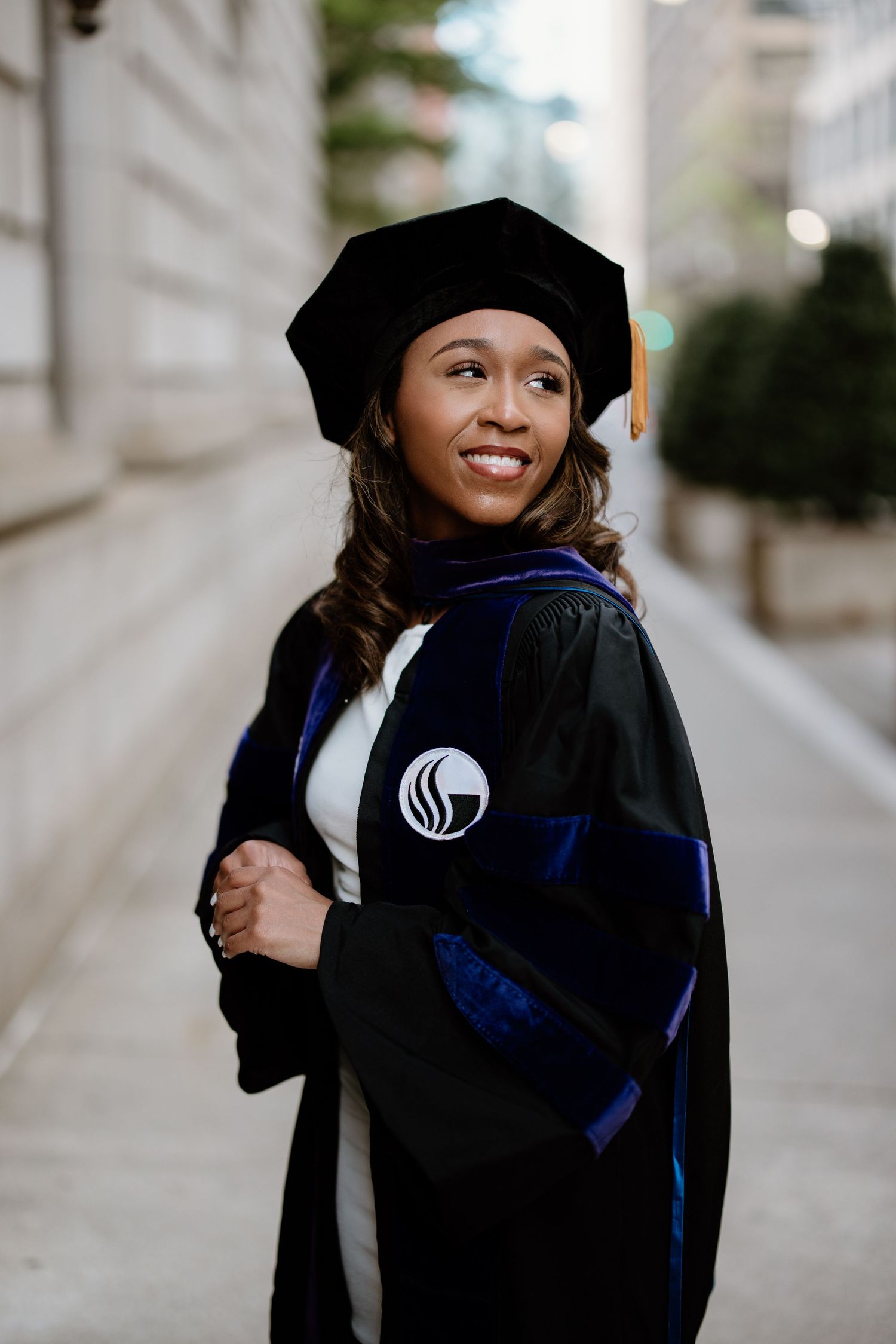 Graduate wearing doctoral regalia with cap and gown smiling on a city street.