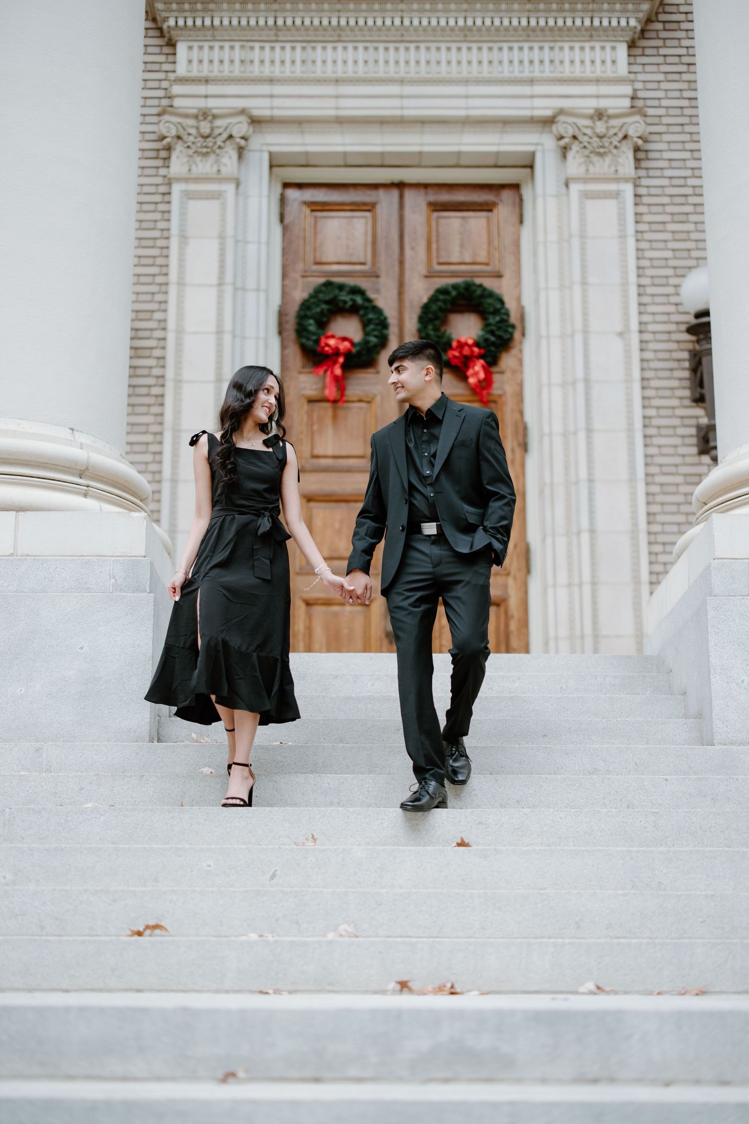 A couple in elegant black attire sitting on grand stone steps with Christmas wreaths decorating wooden doors in the background.