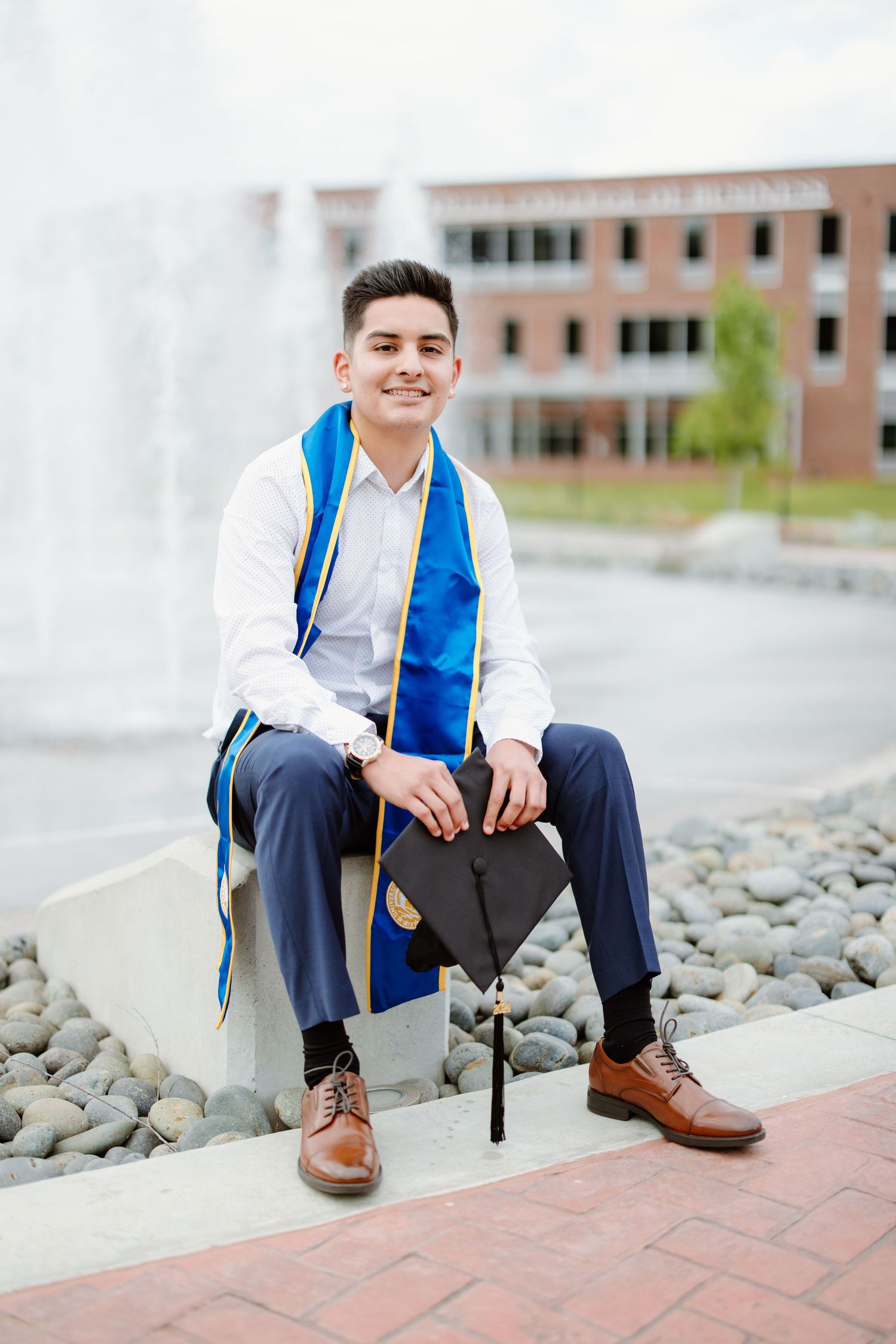 Graduation portrait with blue and white stole sitting on rocks near fountain outside brick university building.