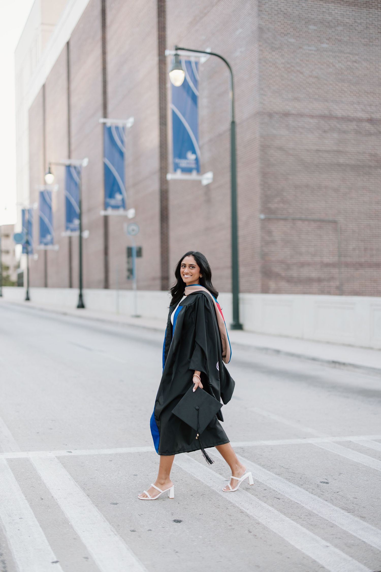 A smiling woman in a black graduation gown and cap walks across a street, celebrating her academic achievement.