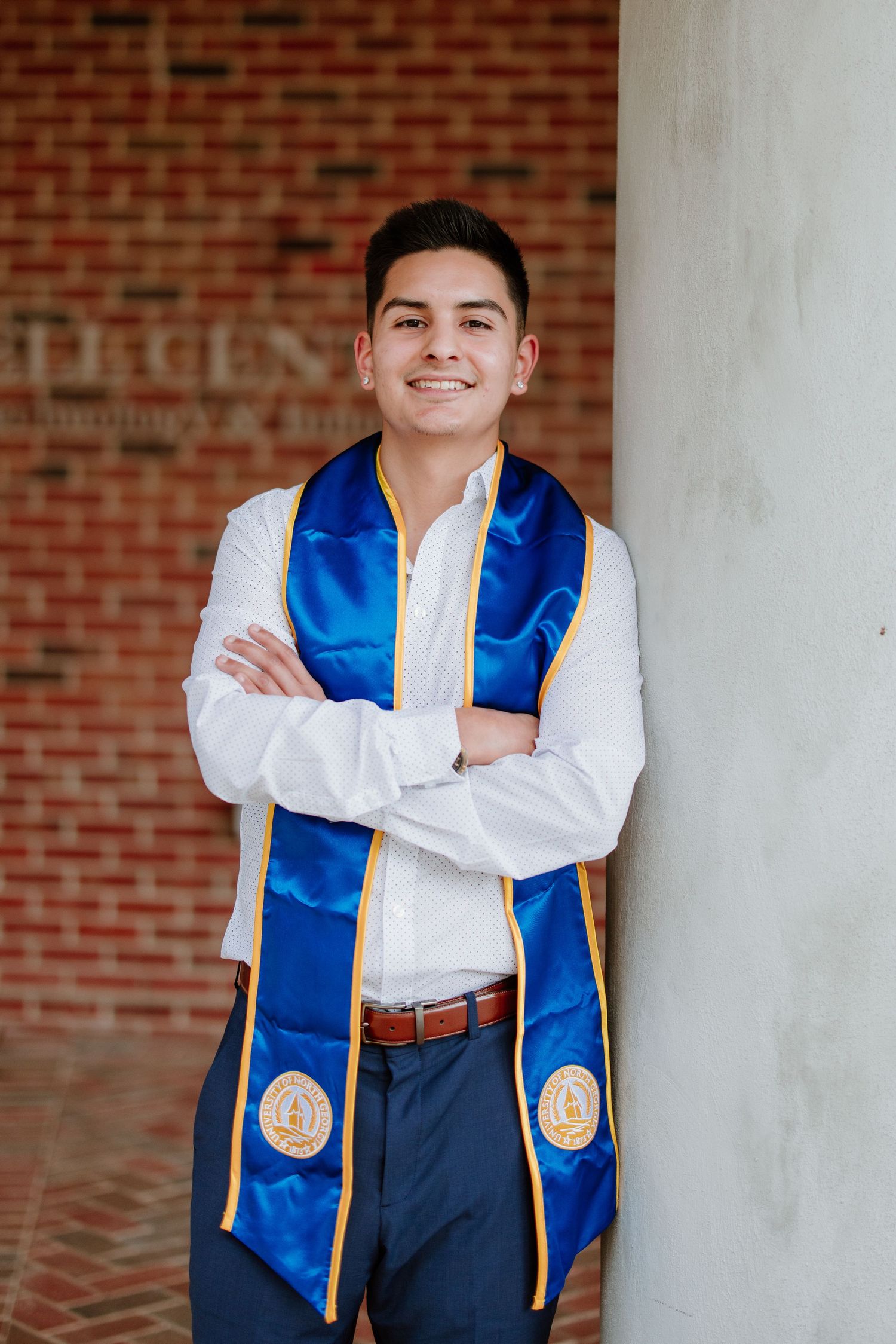 Graduate wearing blue and gold stole poses against brick wall during commencement photo session.