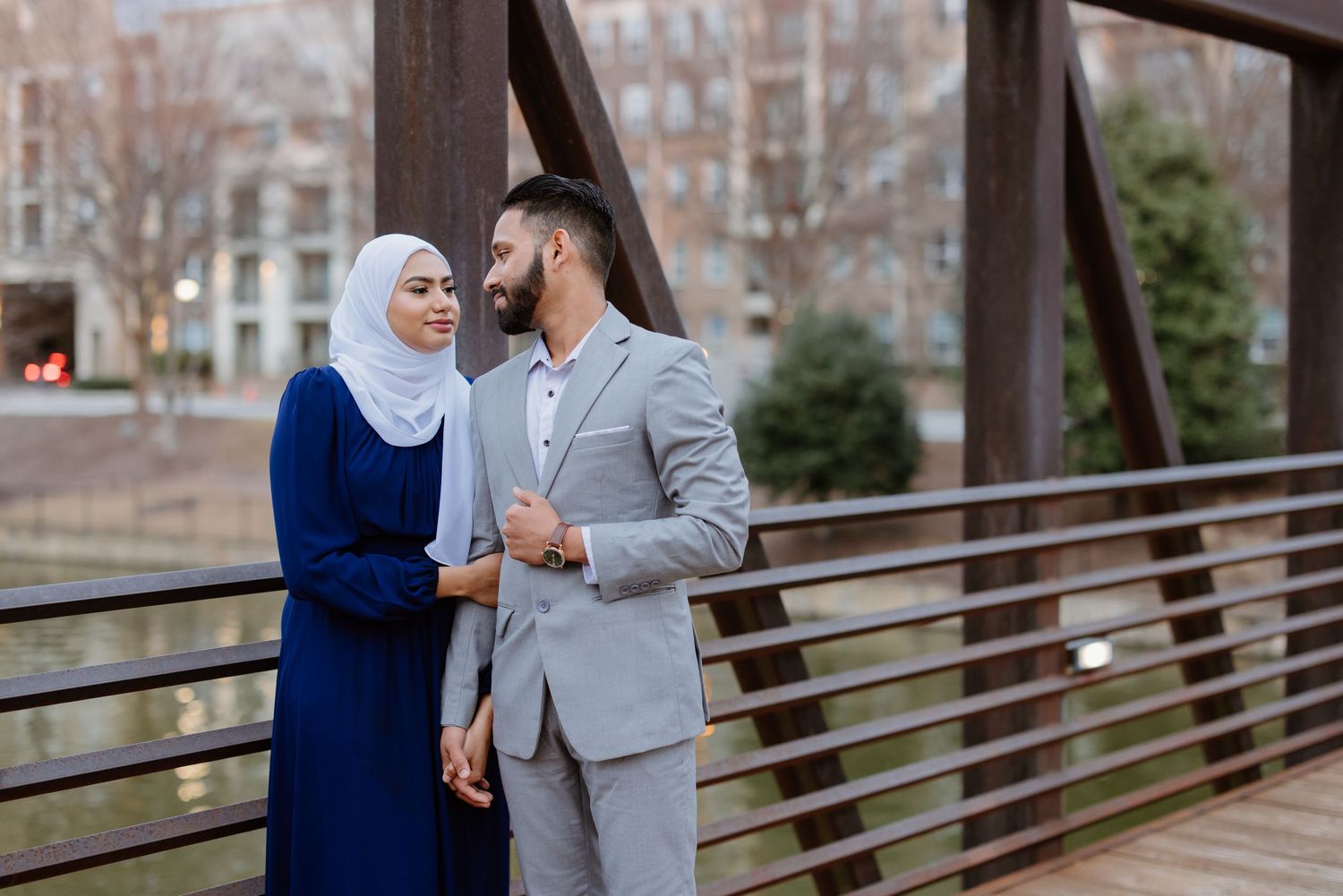 Couple in coordinated formal attire pose together on an outdoor bridge walkway with city buildings in background.