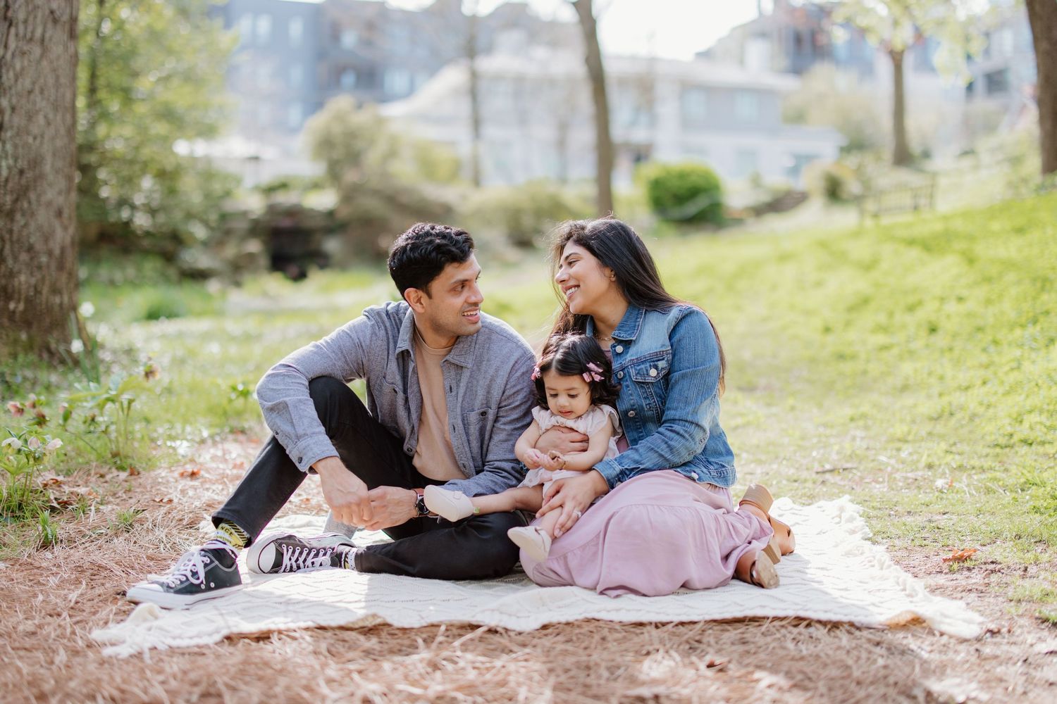 A couple picnicking in a park with their baby, sitting on a white blanket under trees on a sunny day.