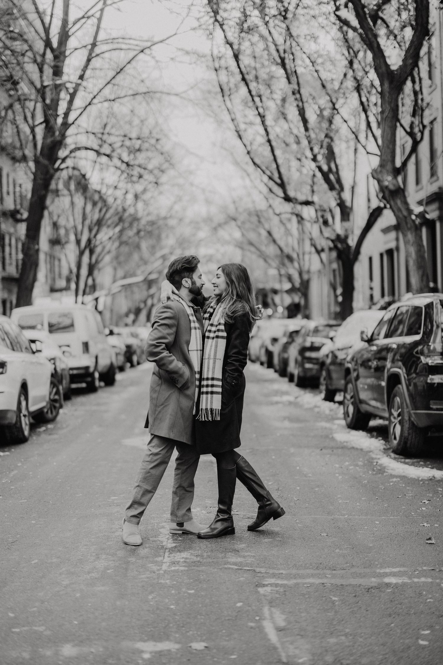A romantic couple embraces in the middle of a tree-lined city street with parked cars on both sides in black and white.