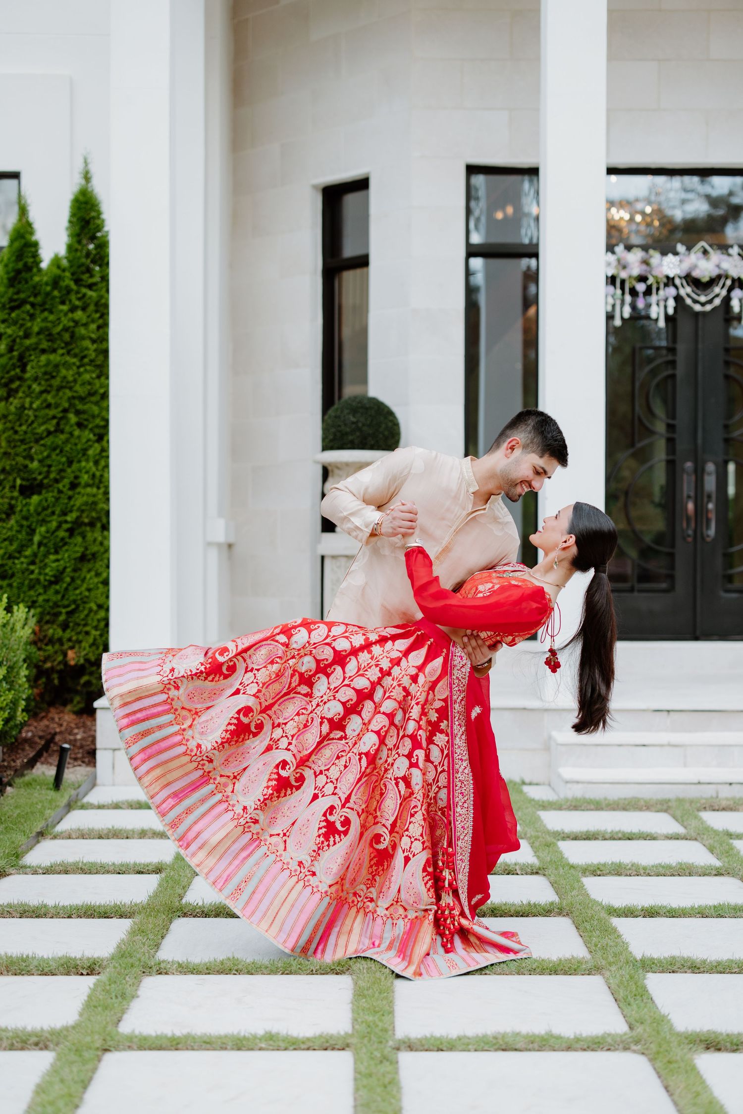 A couple in traditional wedding attire dance gracefully on a paved pathway by a white building with landscaped greenery.