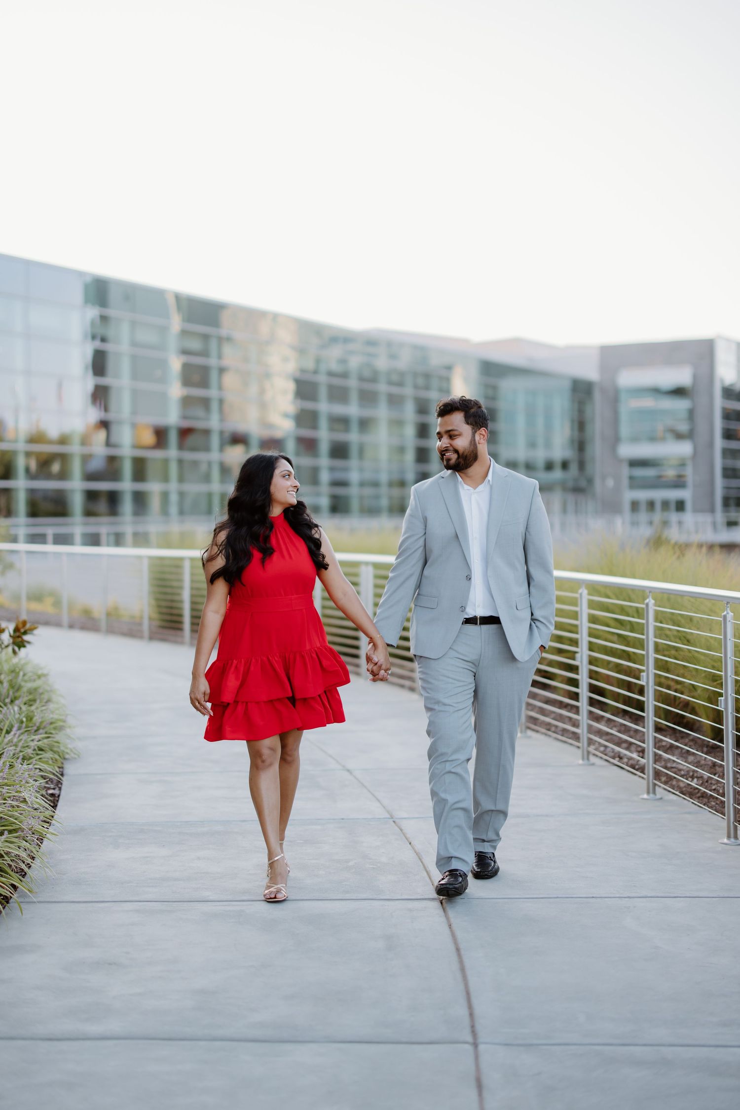 Couple walking together on urban path, with a red dress and gray suit against modern building backdrop.