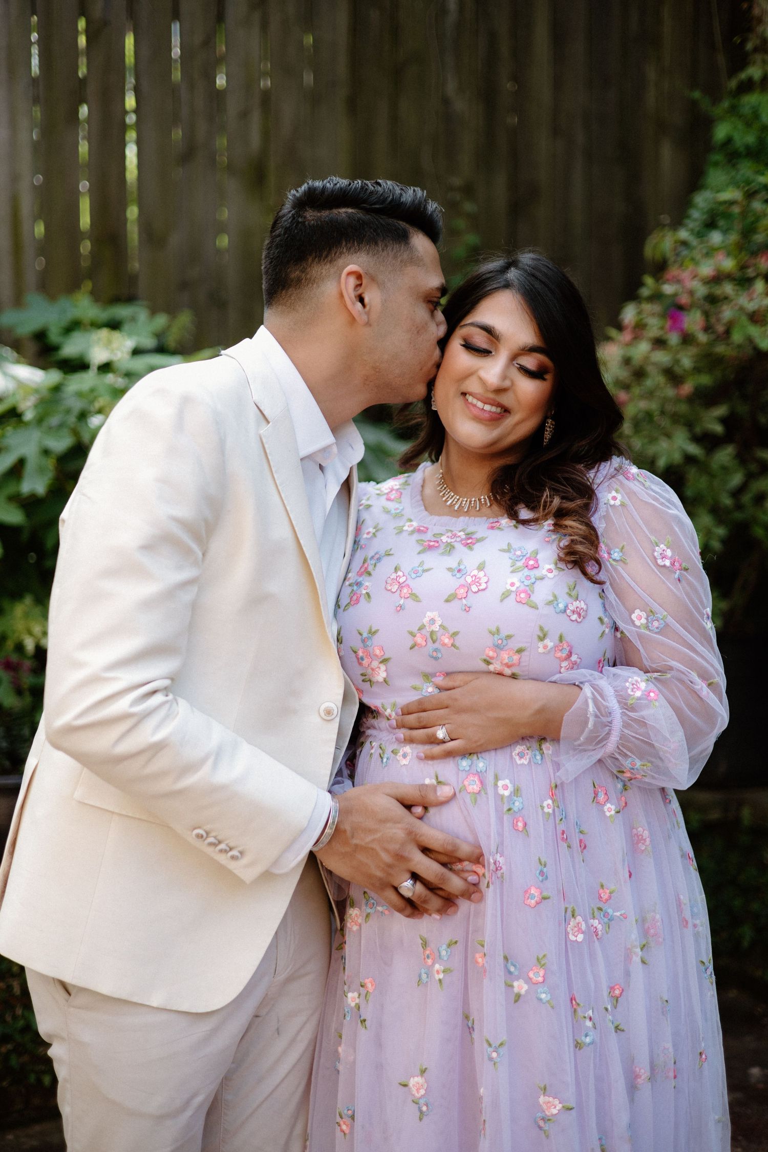 Couple in white and floral pink outfits sharing tender moments in an intimate outdoor maternity photo shoot.