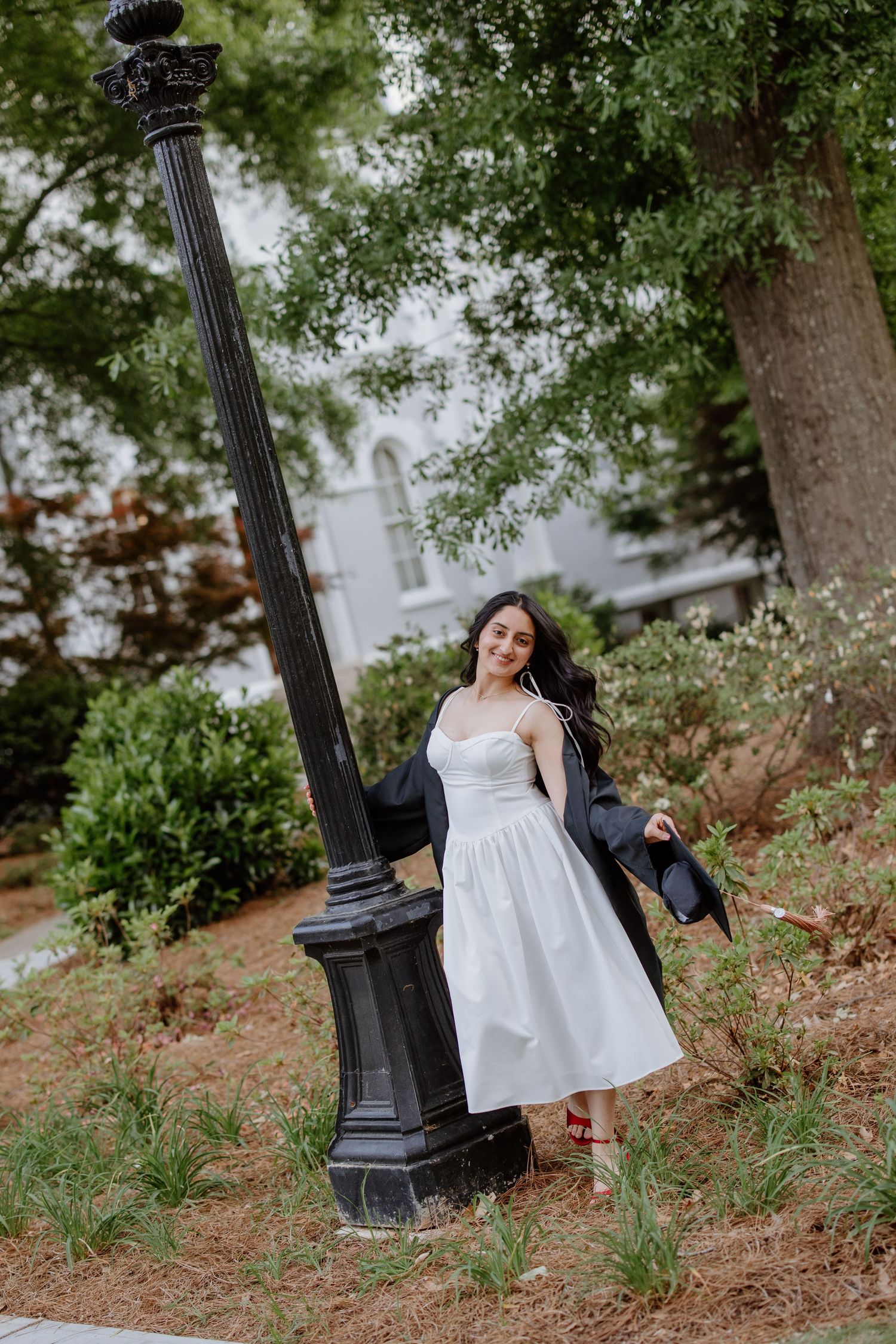 Young woman in white dress and black graduation gown poses by ornate lamp post in lush garden setting.