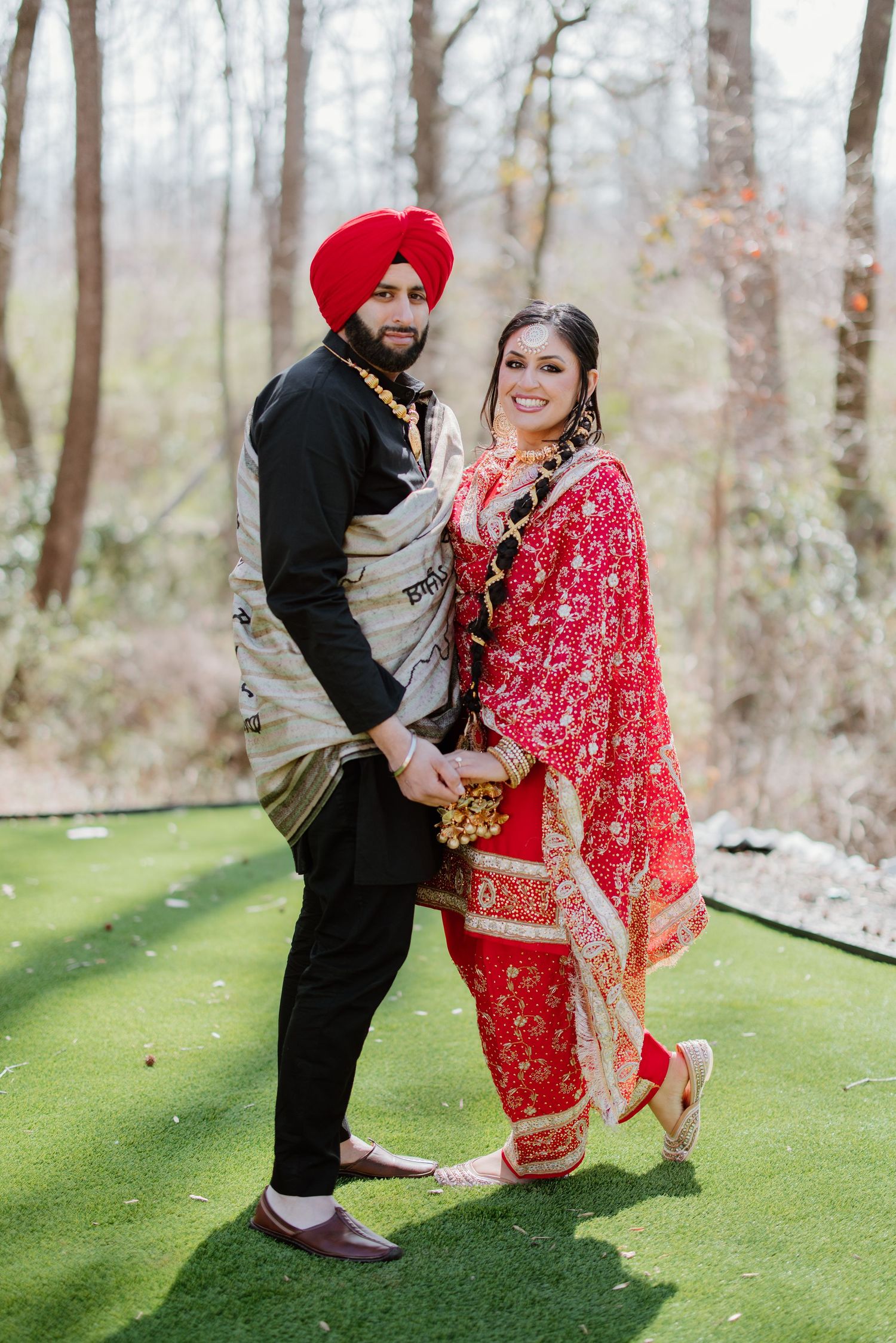 A couple in traditional Punjabi wedding attire poses together on a green surface in a wooded outdoor setting.