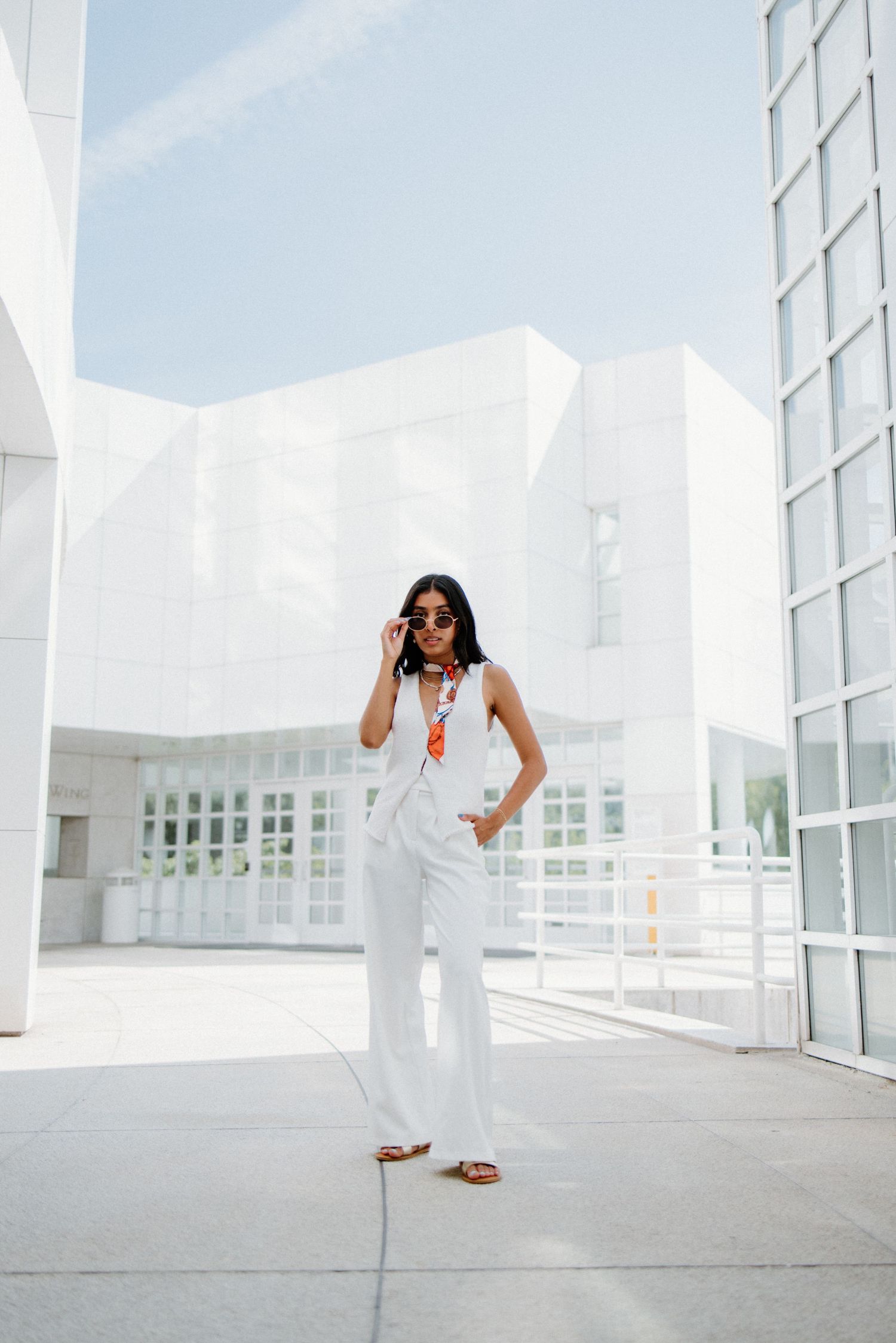 Person in white dress and sunglasses poses confidently in bright minimalist architectural setting with large windows.