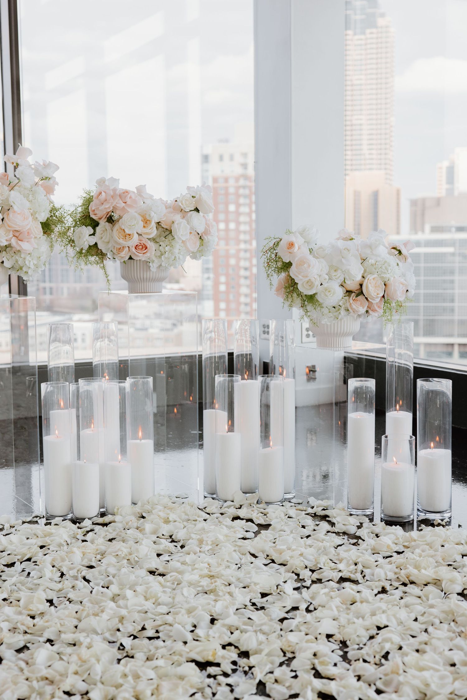 Elegant white wedding reception decor with rose petals and candles against a city skyline view through glass windows.