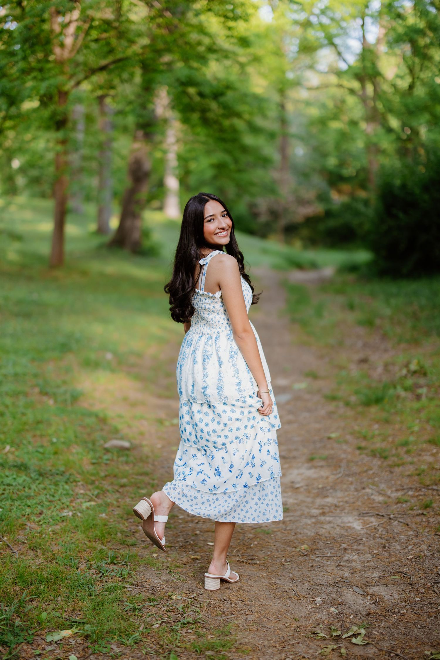 Figure in a white and blue floral midi dress poses gracefully on a wooded path during golden hour.