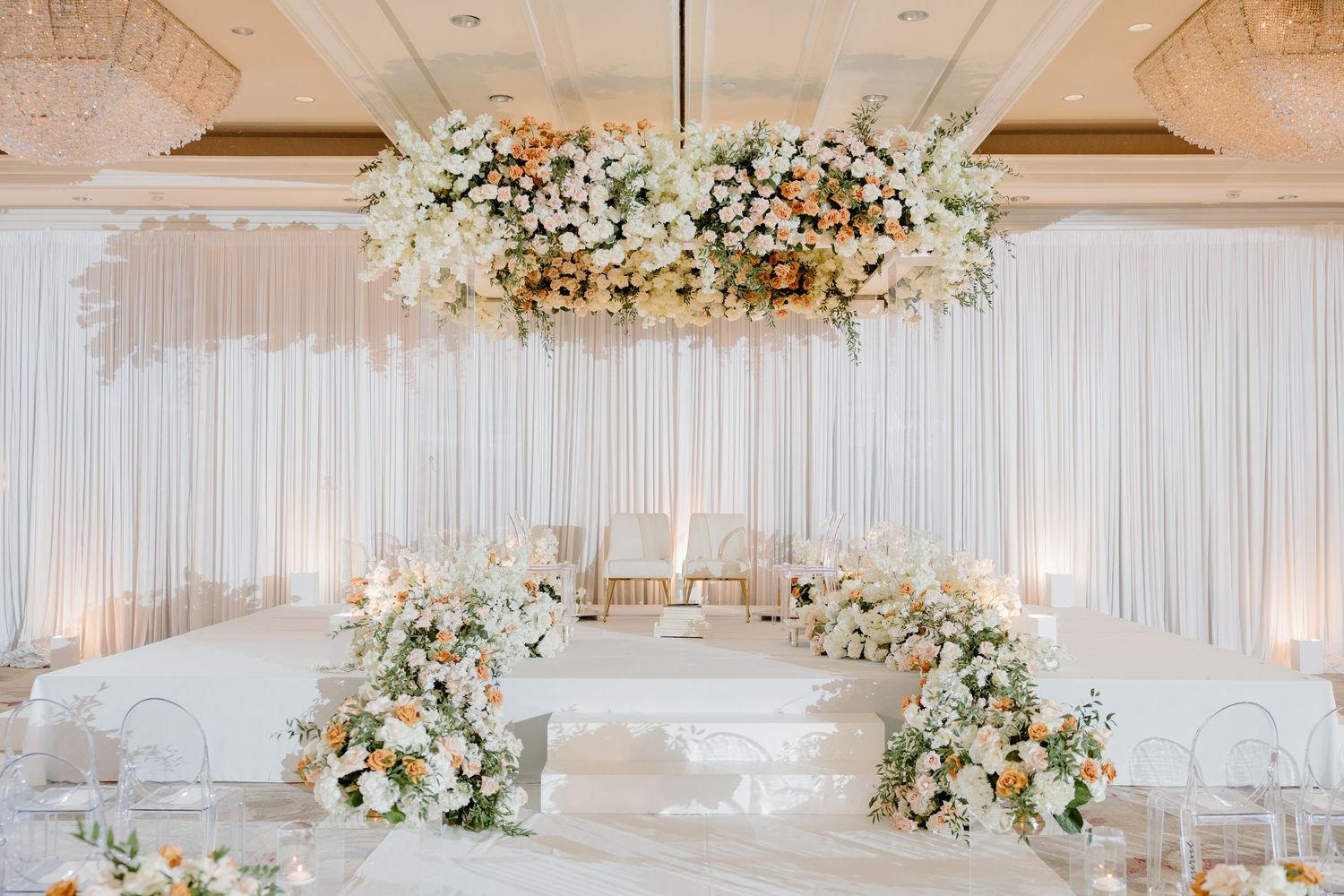 Stunning white draped wedding reception hall with floral chandelier centerpieces and ambient lighting.