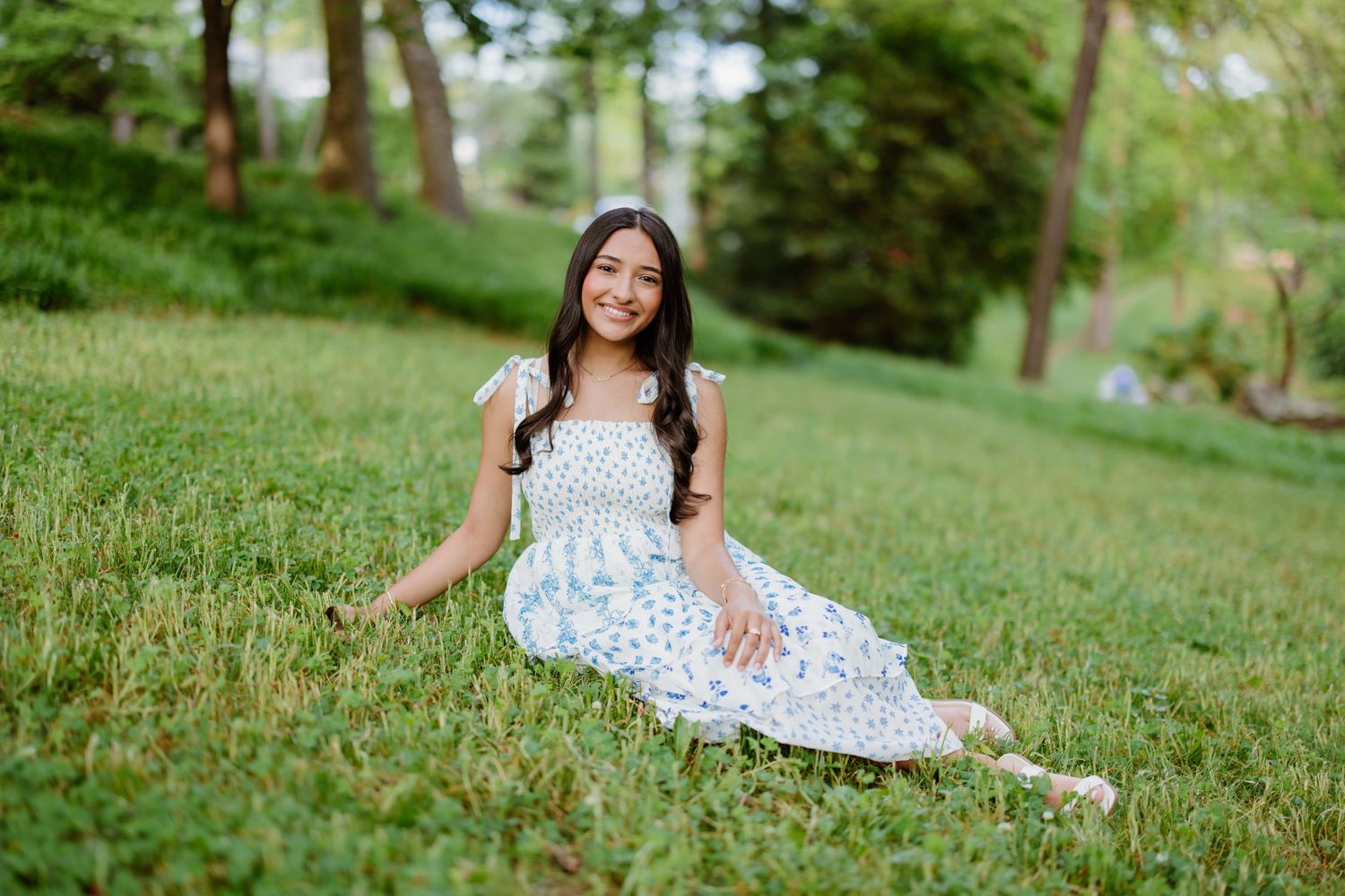 Figure in a white floral dress poses gracefully on a lush green hillside during an outdoor photo session.
