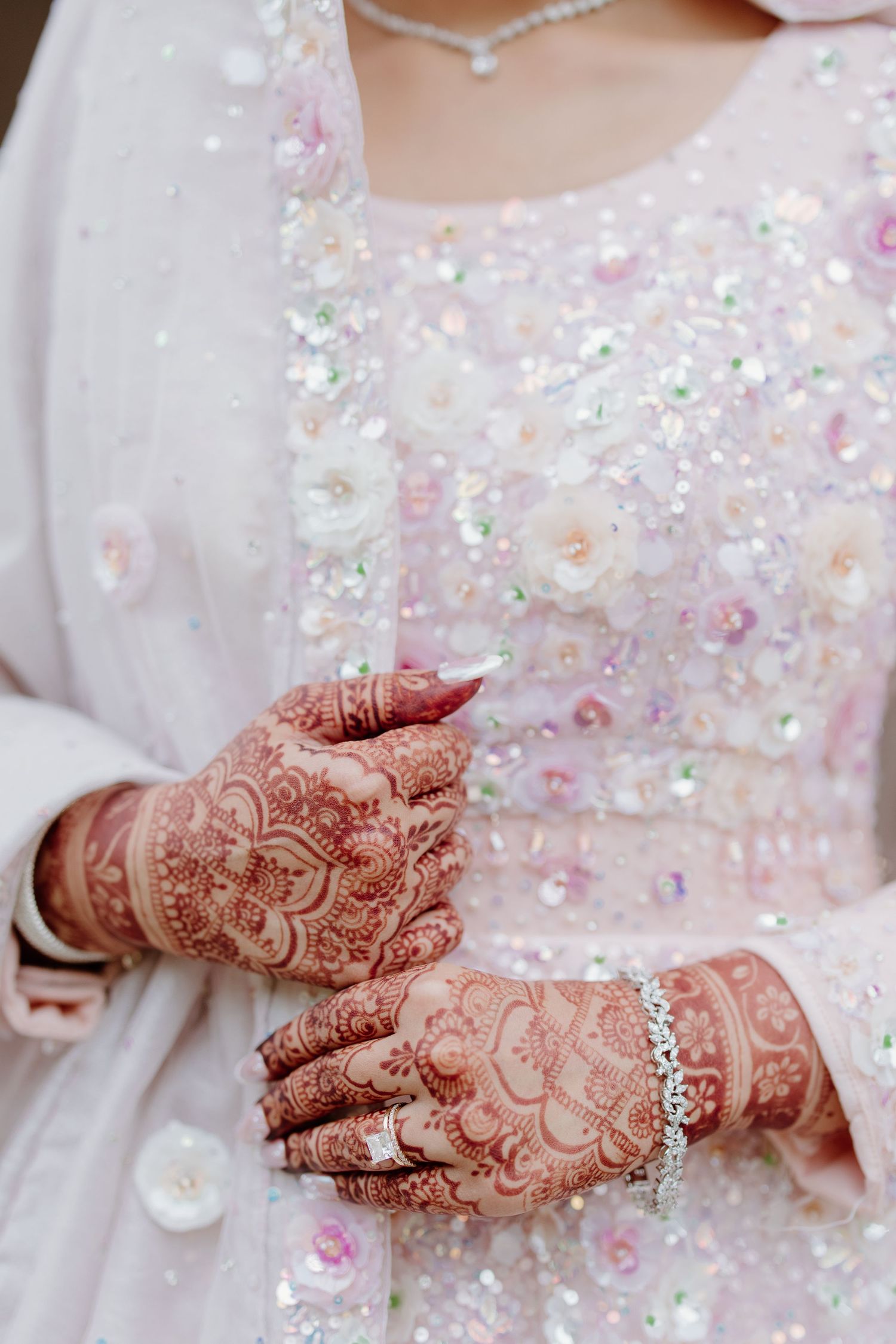 Hands adorned with intricate henna designs resting on a sparkly white sequined dress with pink embellishments and a bracelet.