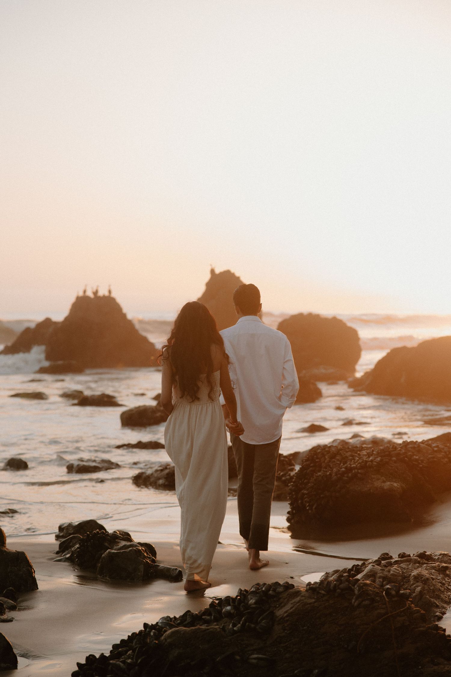 Couple walking hand in hand on a rocky beach at golden sunset, woman in white dress, man in white shirt.