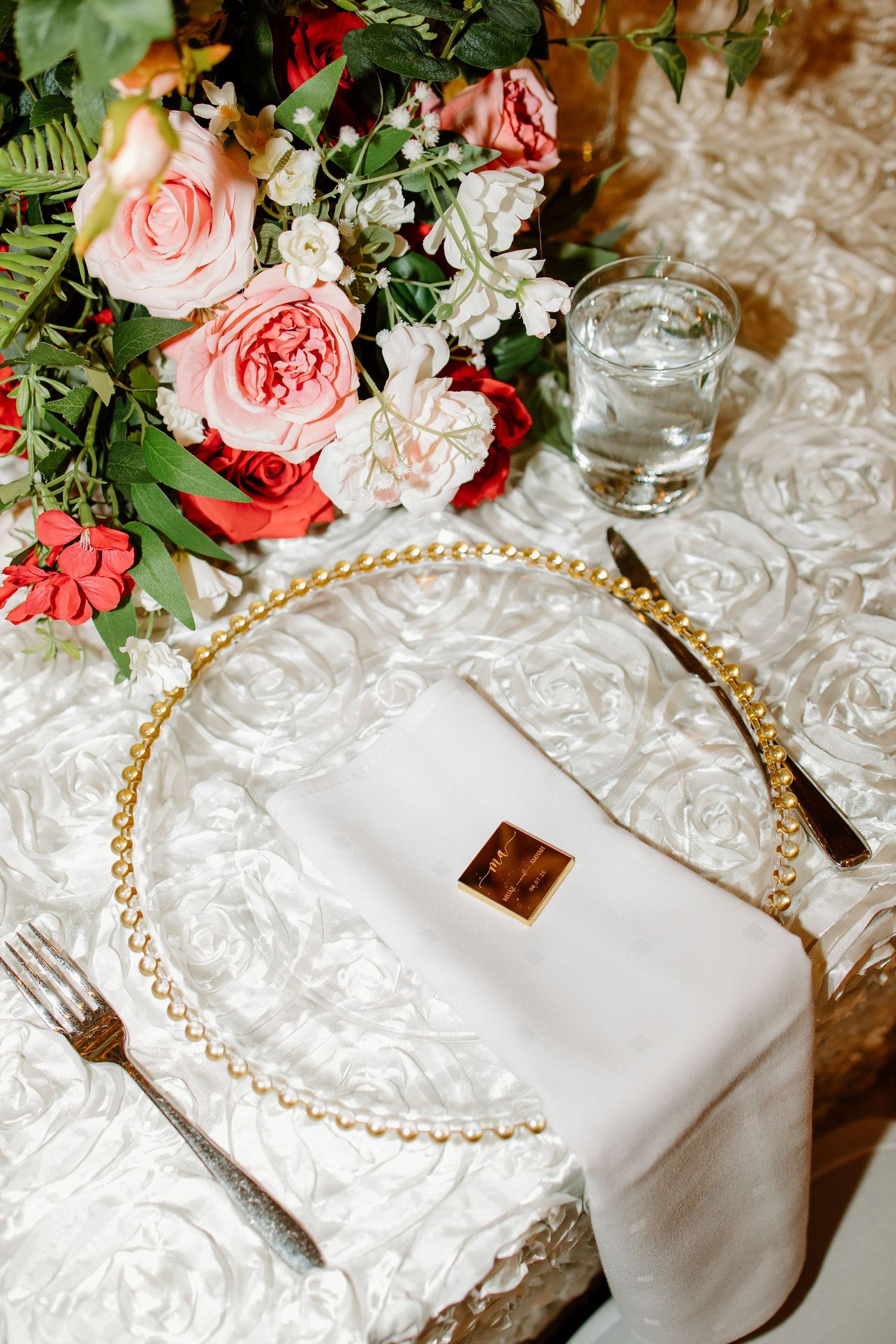 A romantic table setting with pink roses and carnations on white lace linens with gold beaded trim and glass elements.