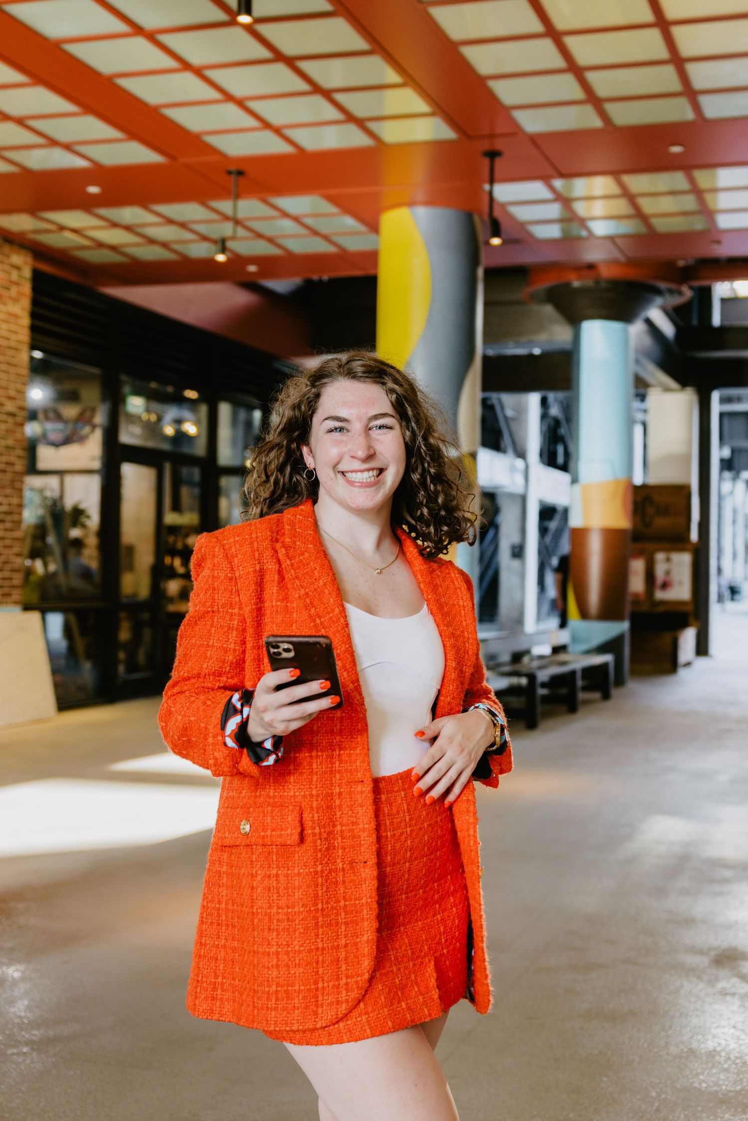 A person wearing a bright orange tweed blazer and skirt set stands in a modern office hallway with brick walls.