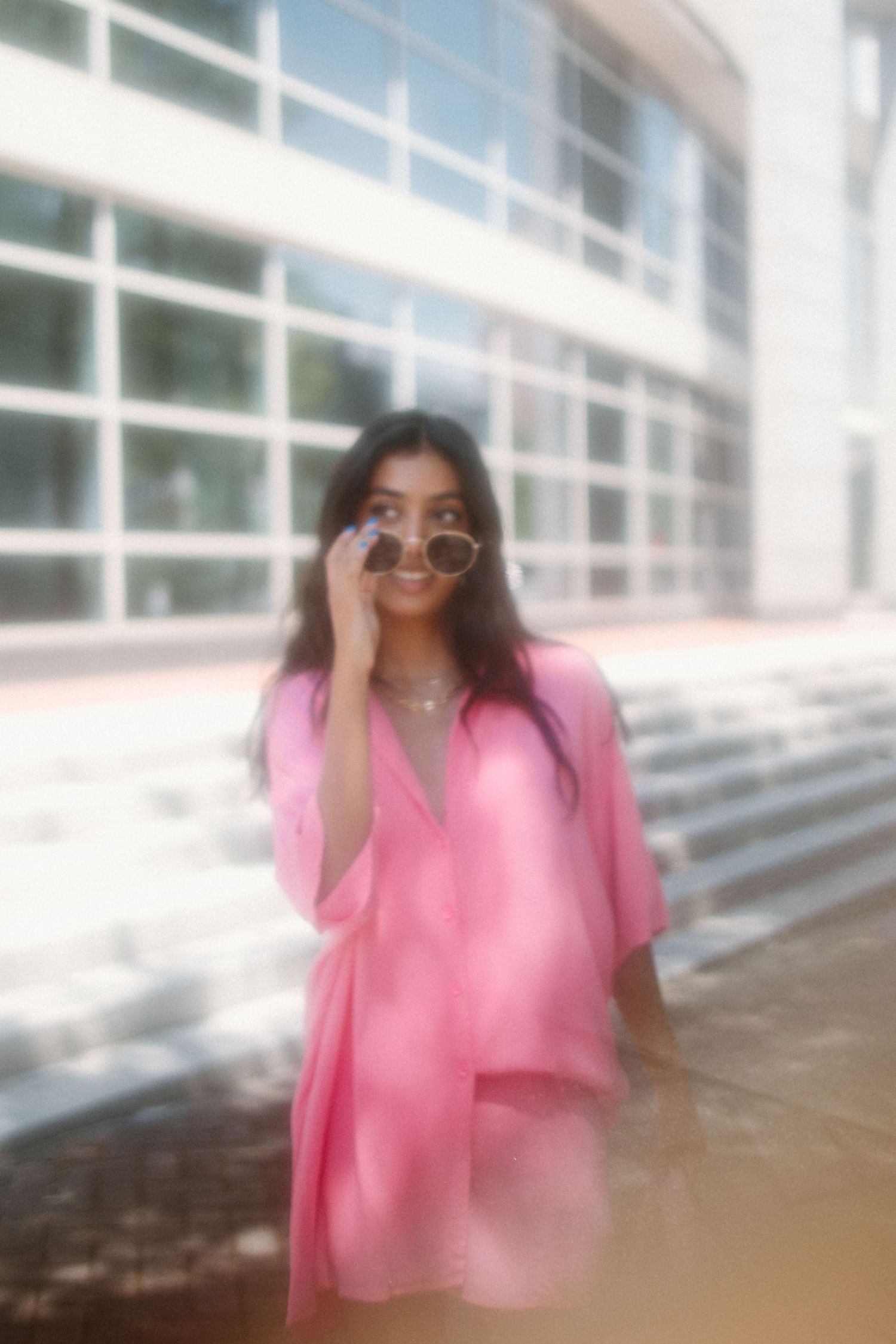 A dreamy soft-focus photo shows someone in a flowing pink oversized shirt and round sunglasses against a modern building.