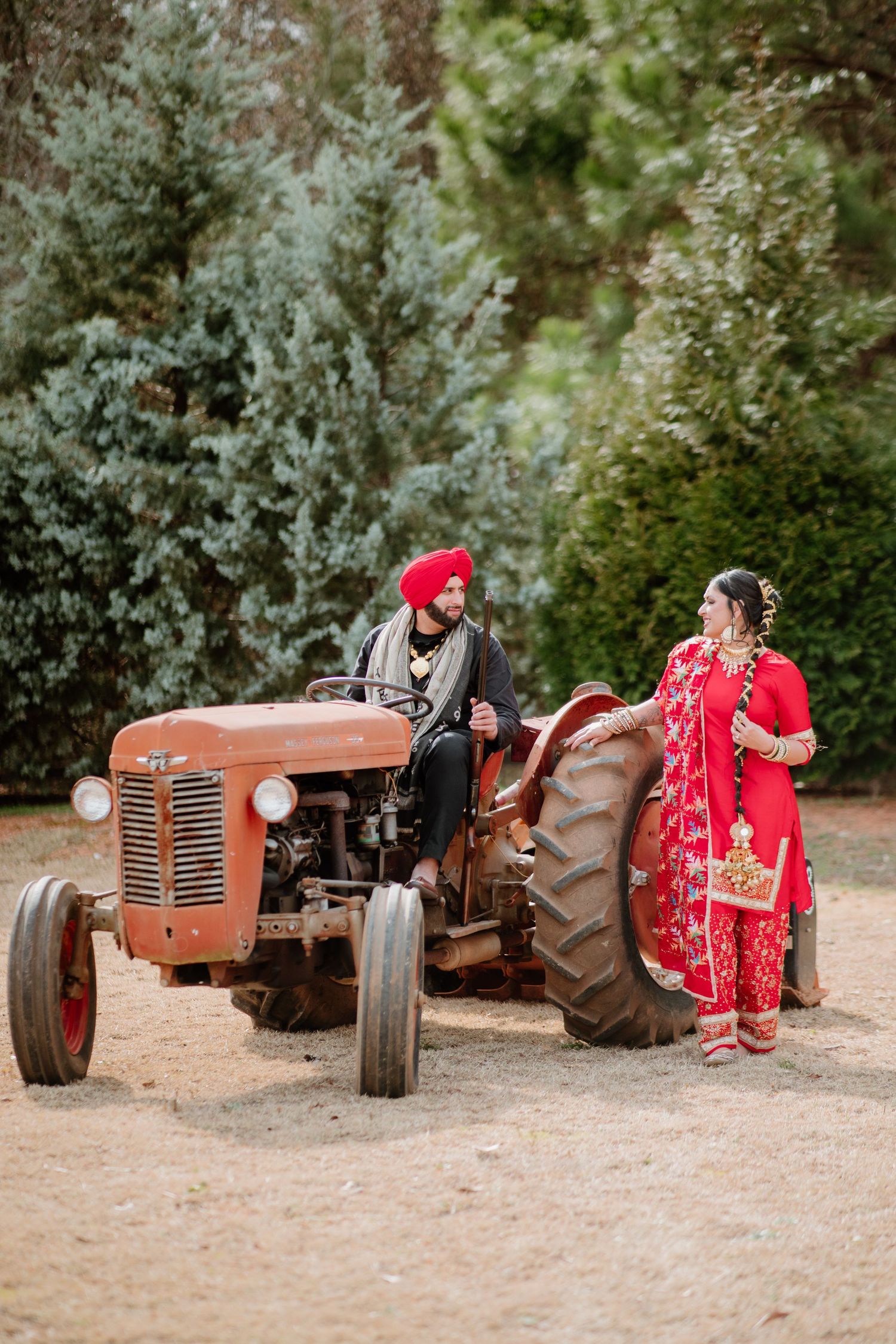 A series of vintage red tractors lined up on a dirt path with someone in a red dress posing beside them.