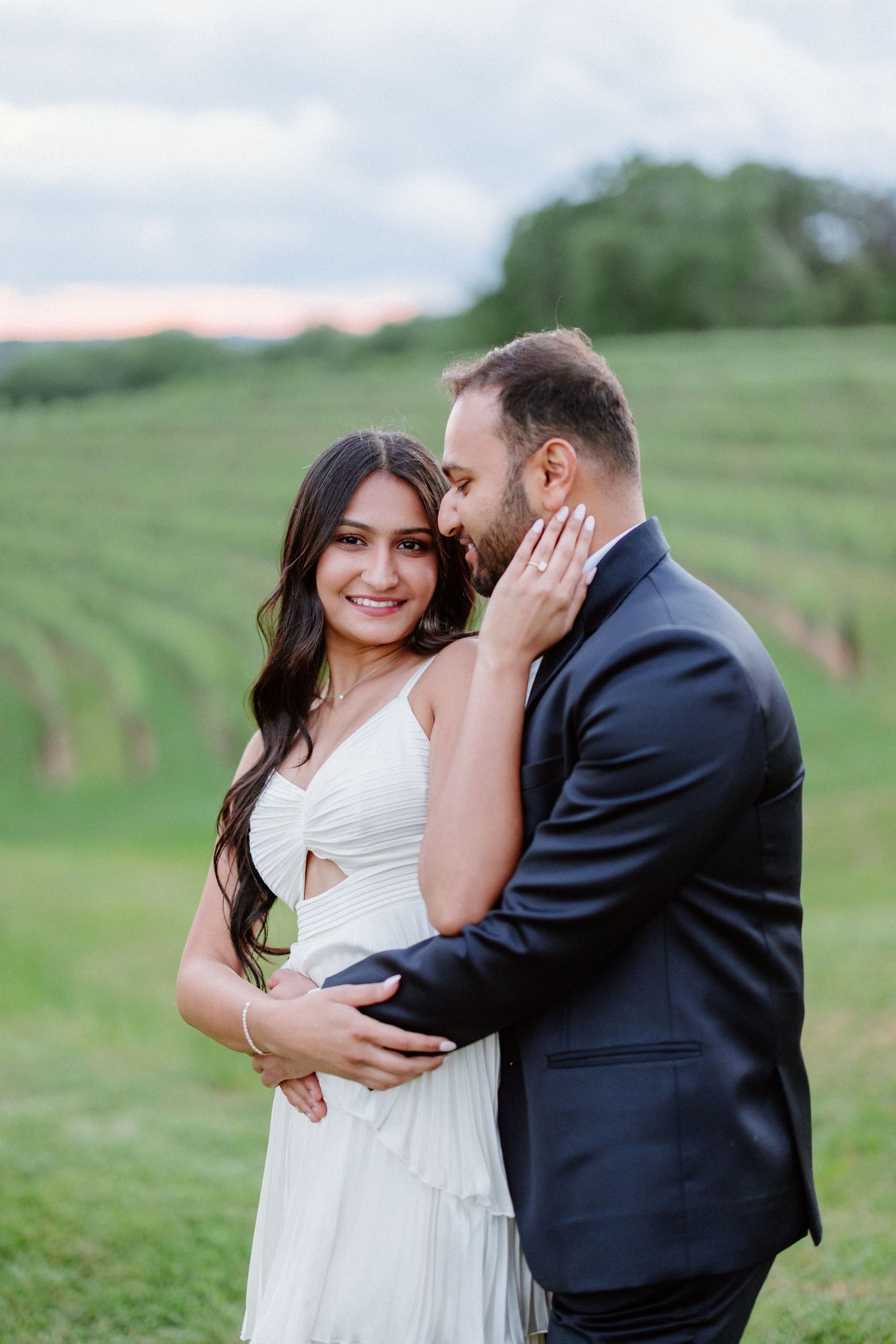 A romantic couple embraces on a lush green hillside during a sunset photo session at an outdoor venue.