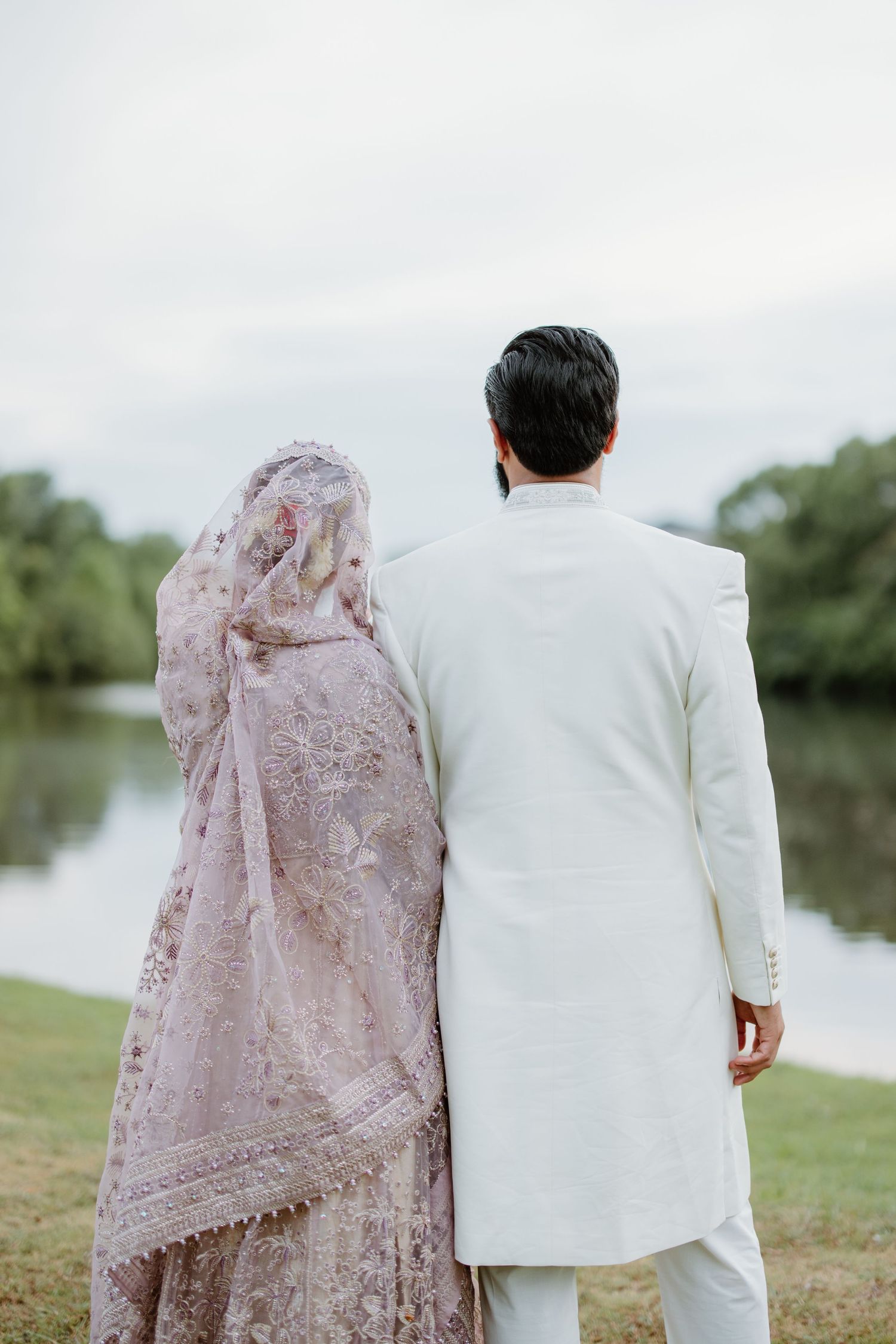 A wedding couple stands together by a lake, with the person wearing a delicate pink lace dress and their partner in white.