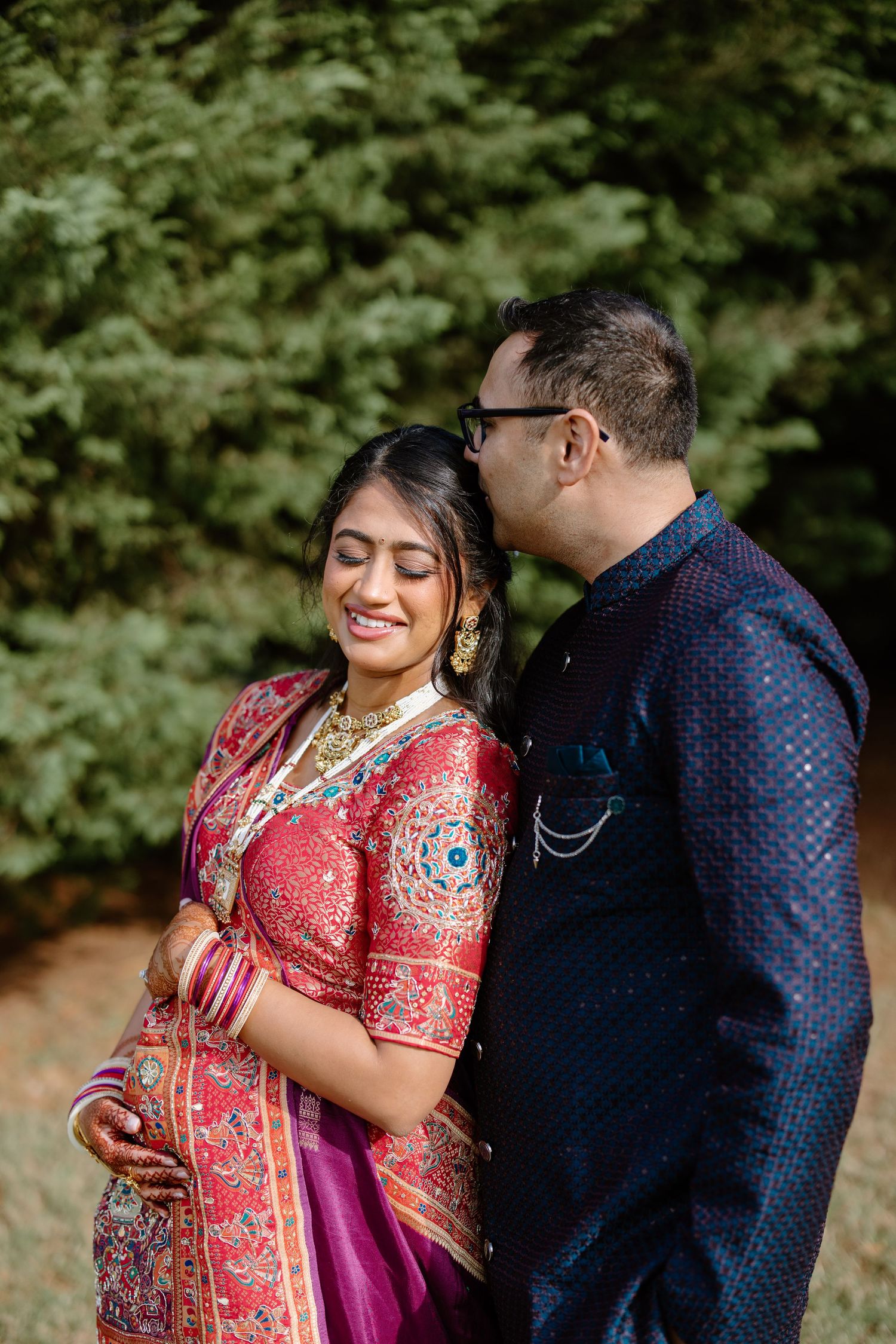 A couple in traditional South Asian attire share an intimate moment outdoors, with the woman wearing a red embroidered sari.