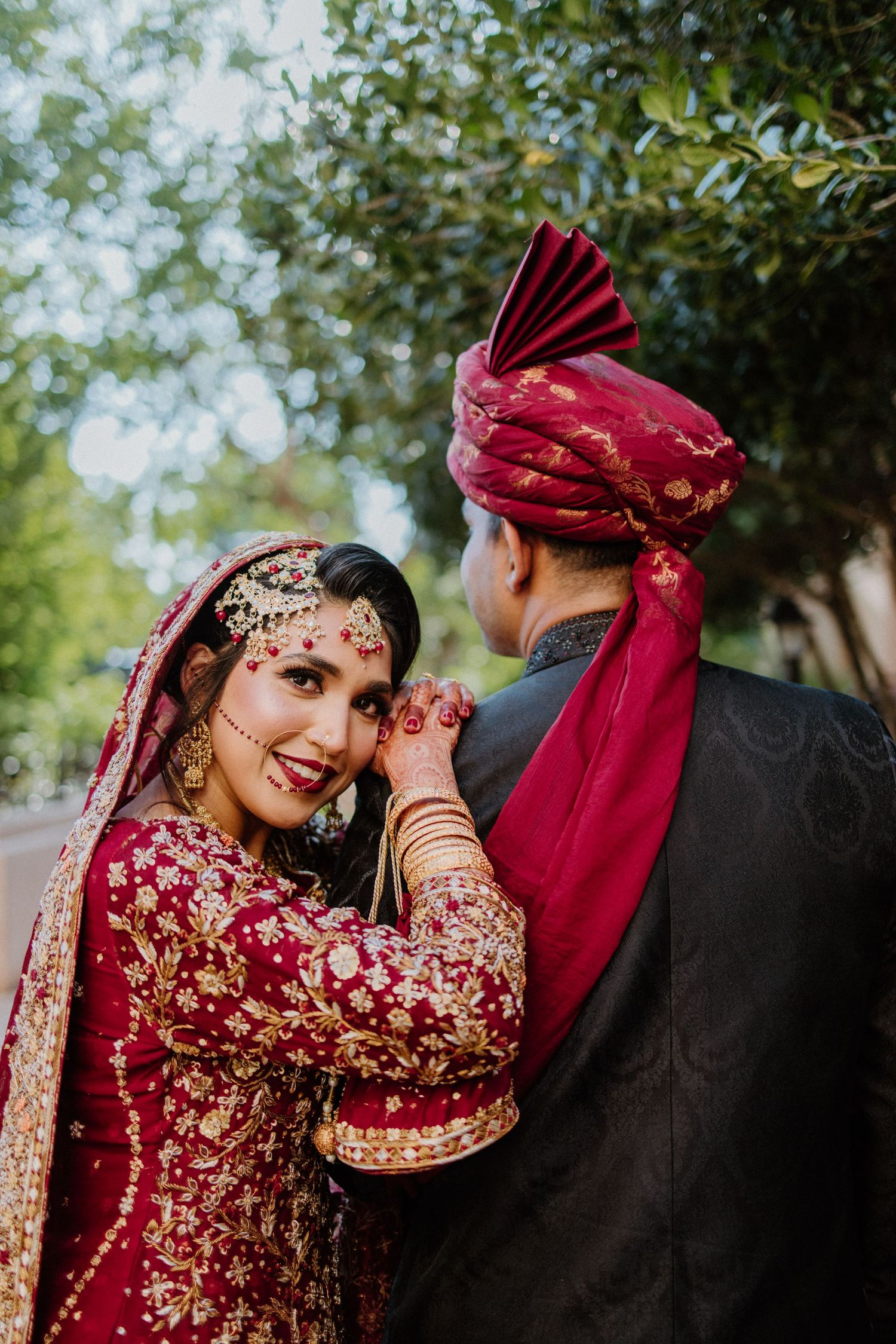 Bride in red and gold embroidered South Asian wedding attire poses with partner in outdoor garden setting.