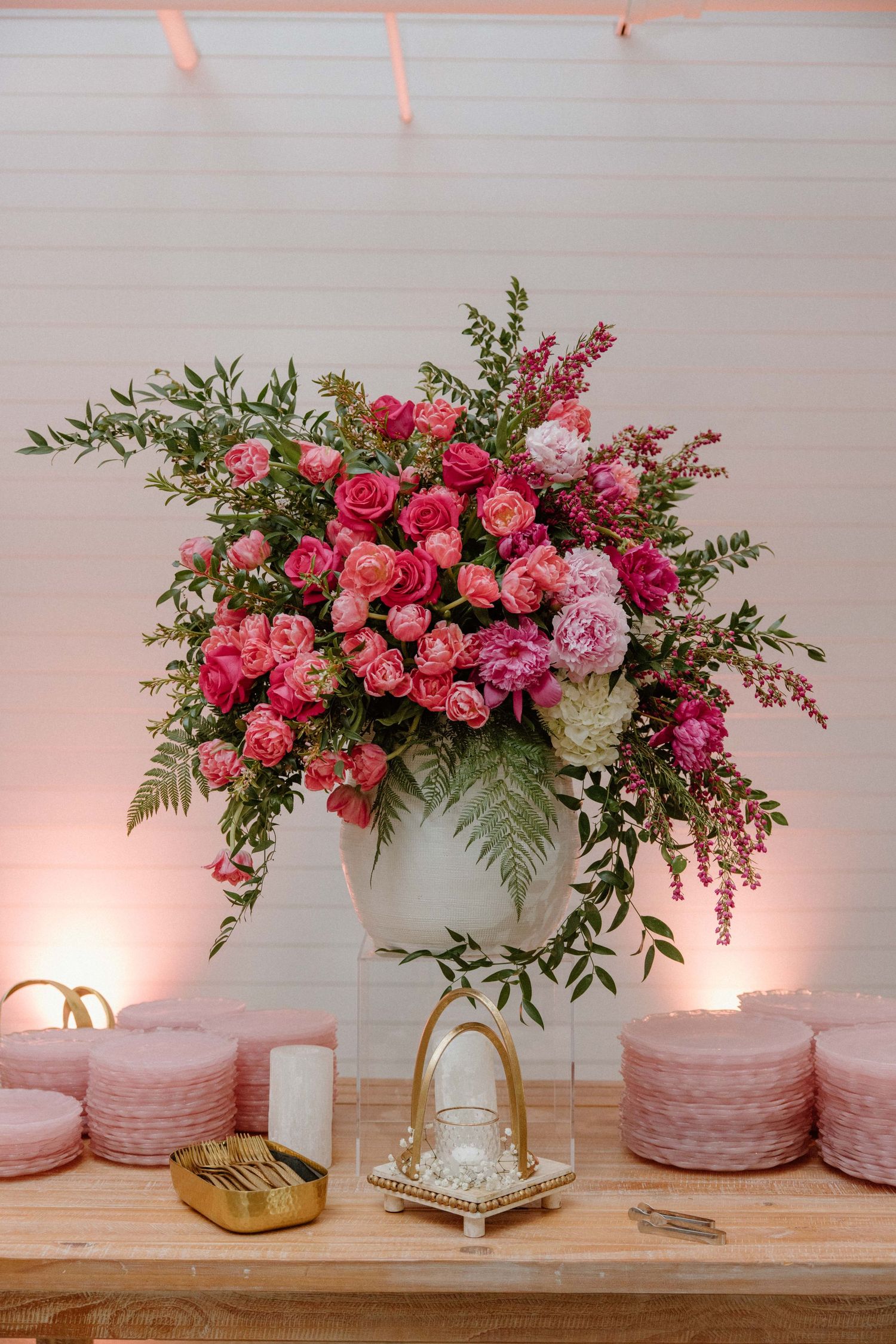 Elegant pink rose floral centerpiece in white vase with gold stand on wooden table with blush pink decor elements.
