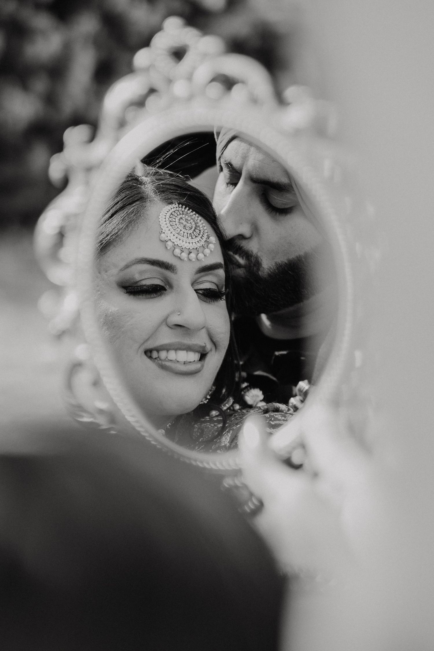 Black and white reflection in ornate mirror shows intimate couple with traditional Indian bridal jewelry and makeup.