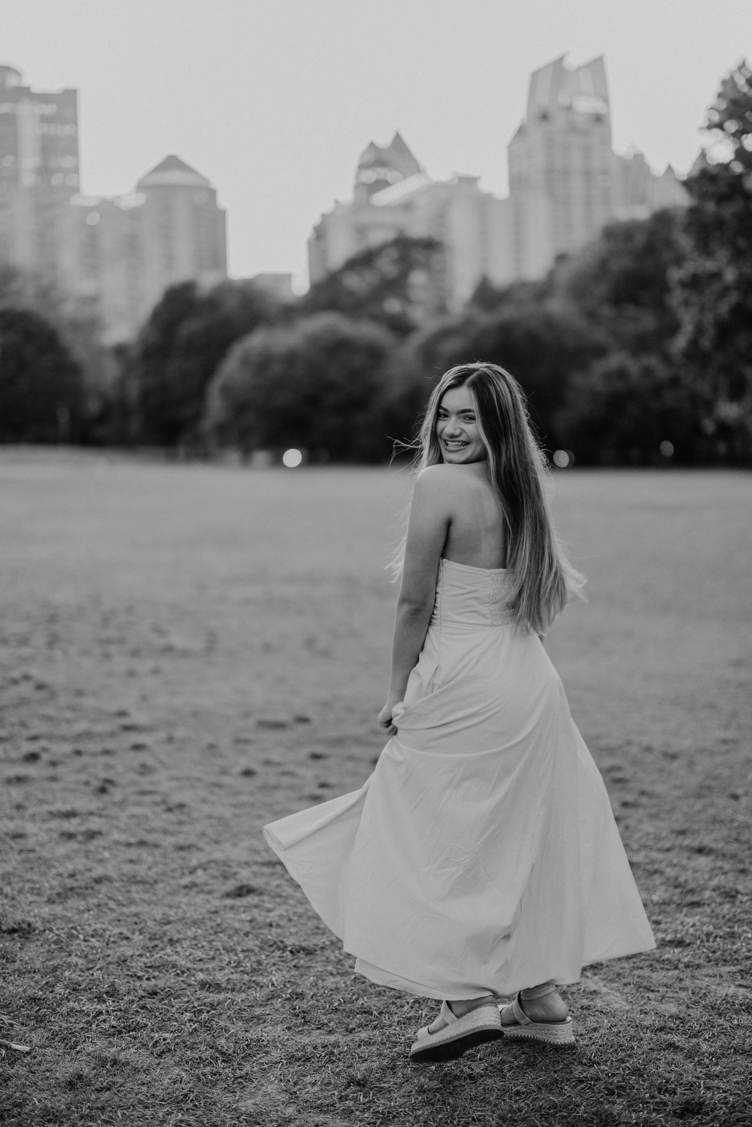 Black and white portrait of a person in a flowing white dress twirling in a city park with skyscrapers in the background.